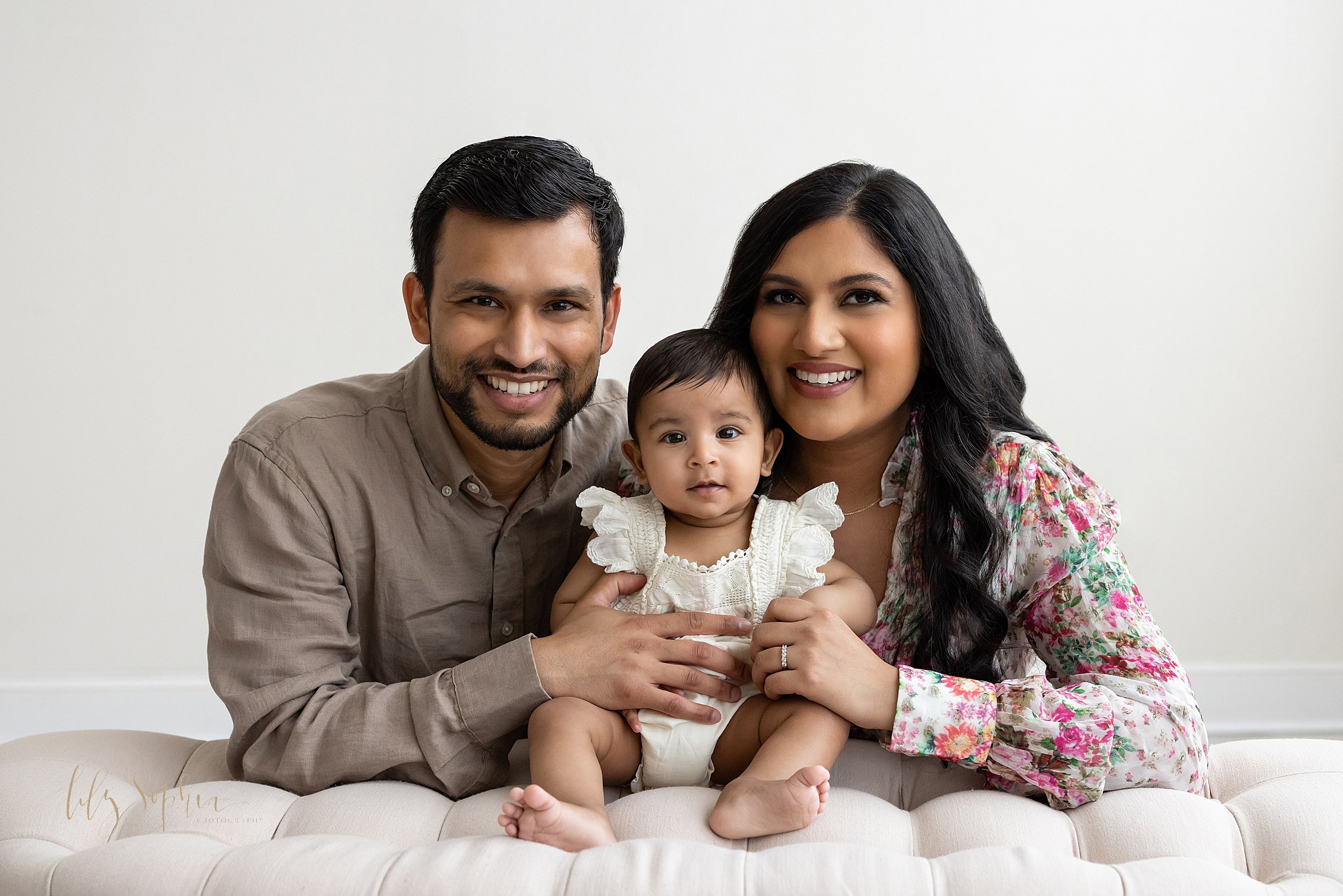  Family milestone portrait with an Indian mom and dad kneeling behind a tufted ottoman as their baby girl sits between them taken in a natural light studio near Buckhead in Atlanta. 