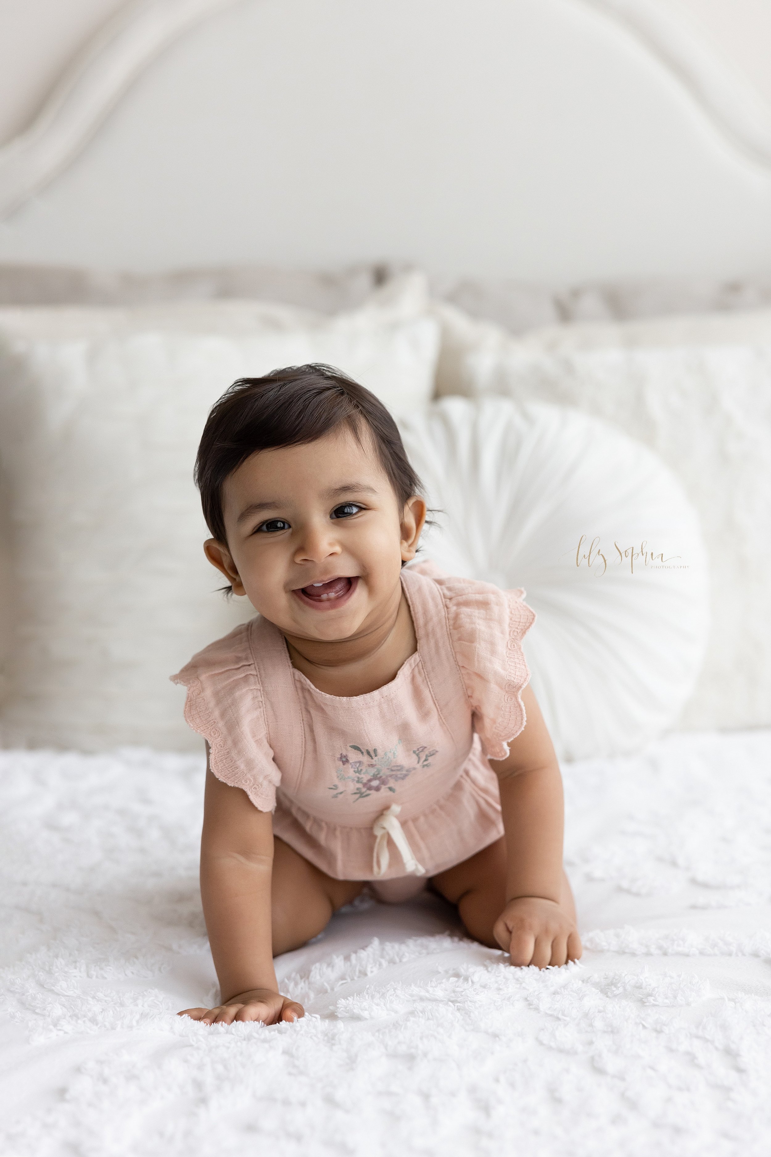  Close-up first birthday portrait of a smiling one year old Indian girl as she crawls on a bed showing her baby teeth taken near Poncey Highlands in Atlanta in a studio using natural light. 