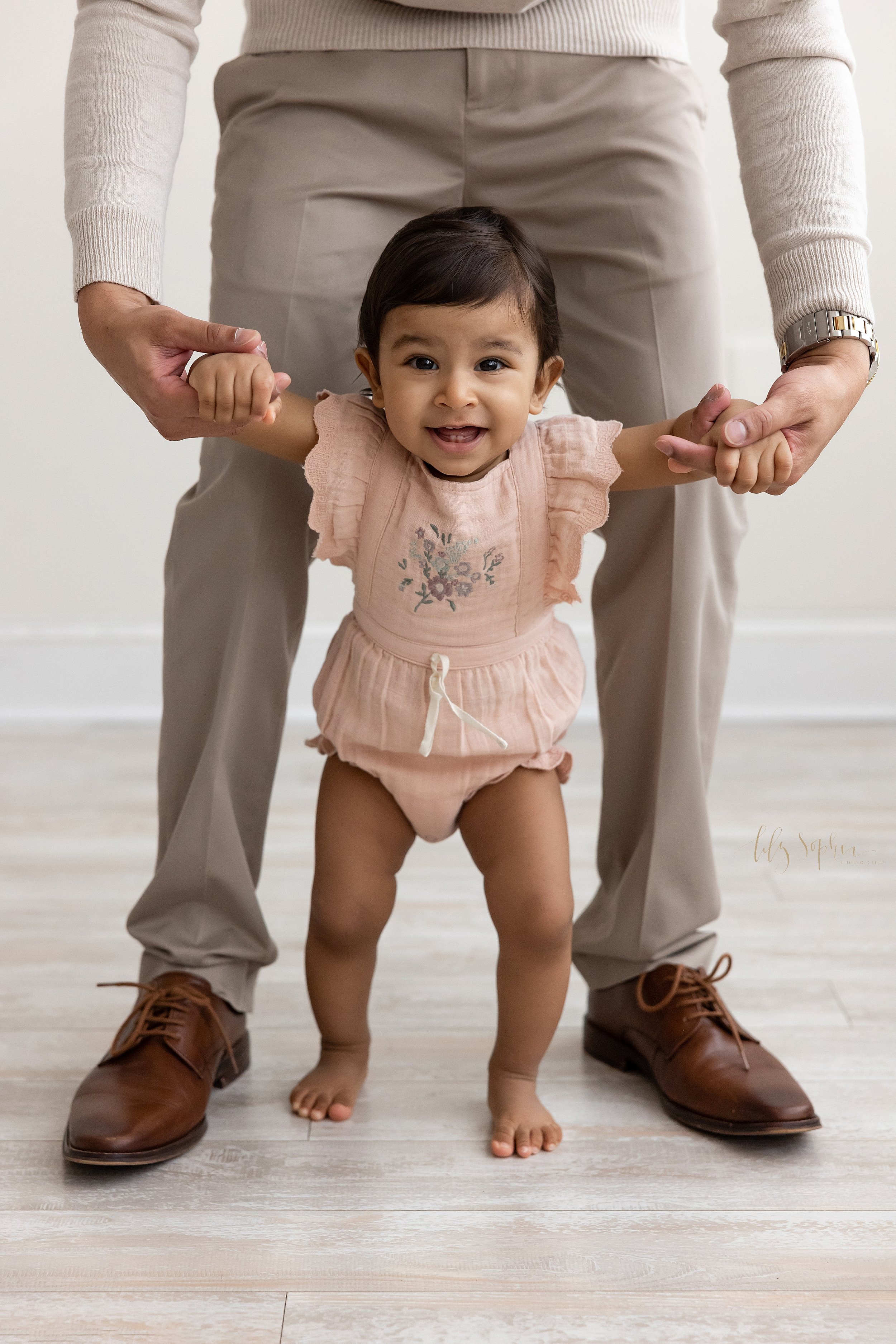  First birthday portrait of a one year old little girl walking barefoot in a natural light studio while holding onto her father’s index fingers as he walks behind her taken near Midtown in Atlanta, Georgia. 