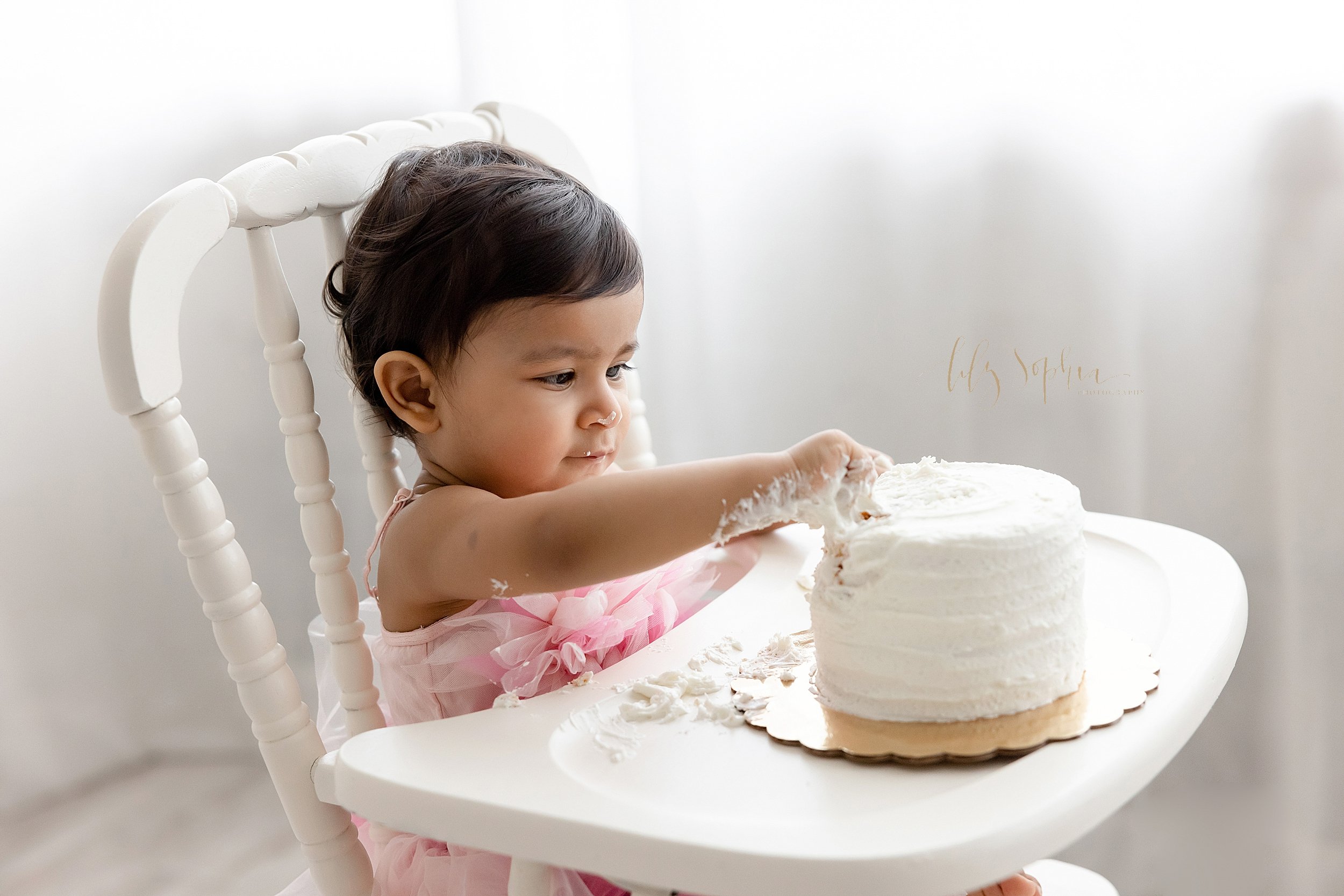  First birthday smash cake determination is captured in this picture of a one year old little Indian girl as she sits in an antique highchair in a studio near Decatur that uses natural light. 