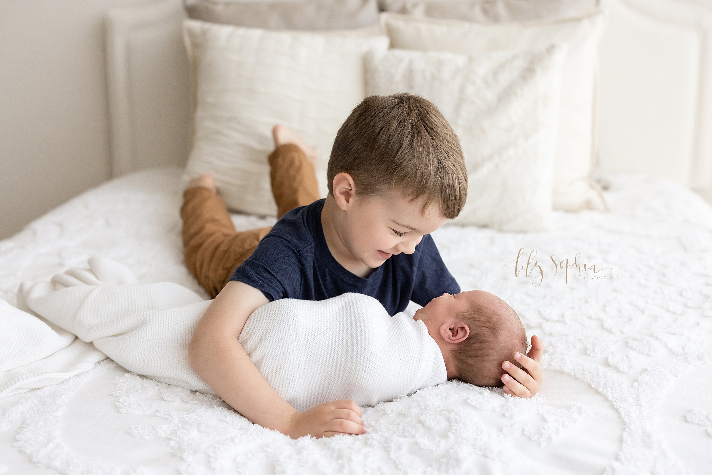  Newborn family photo session with a big brother lying on his stomach on a bed holding the head of his newborn baby brother as he admires him taken near Cummings in Atlanta in a natural light studio. 
