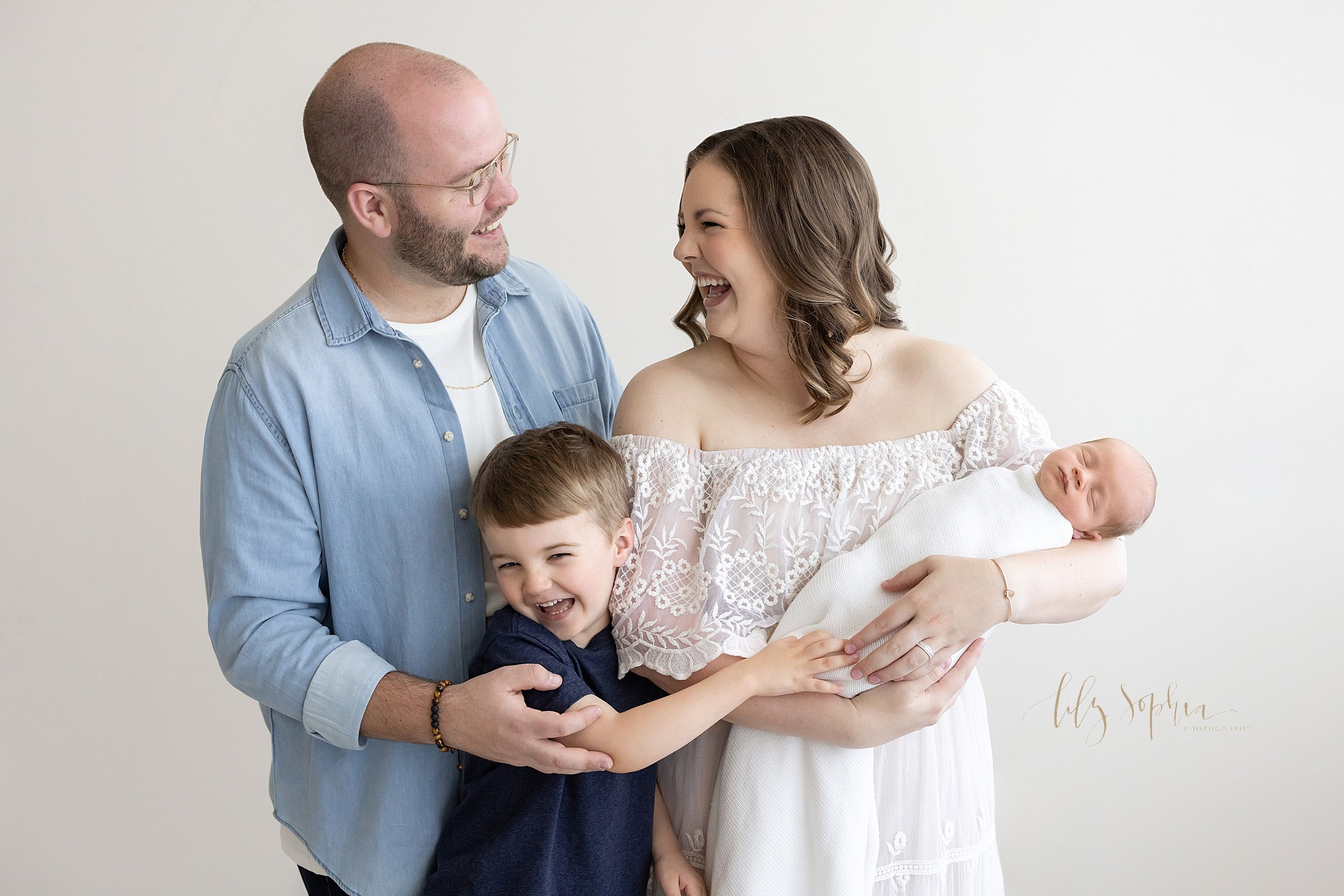  Newborn family photo of a mother cradling her peacefully sleeping newborn baby boy in her arms with her son standing beside her and holding her right arm as her husband stands behind her son with his hand on his son’s shoulder as the family laughs t