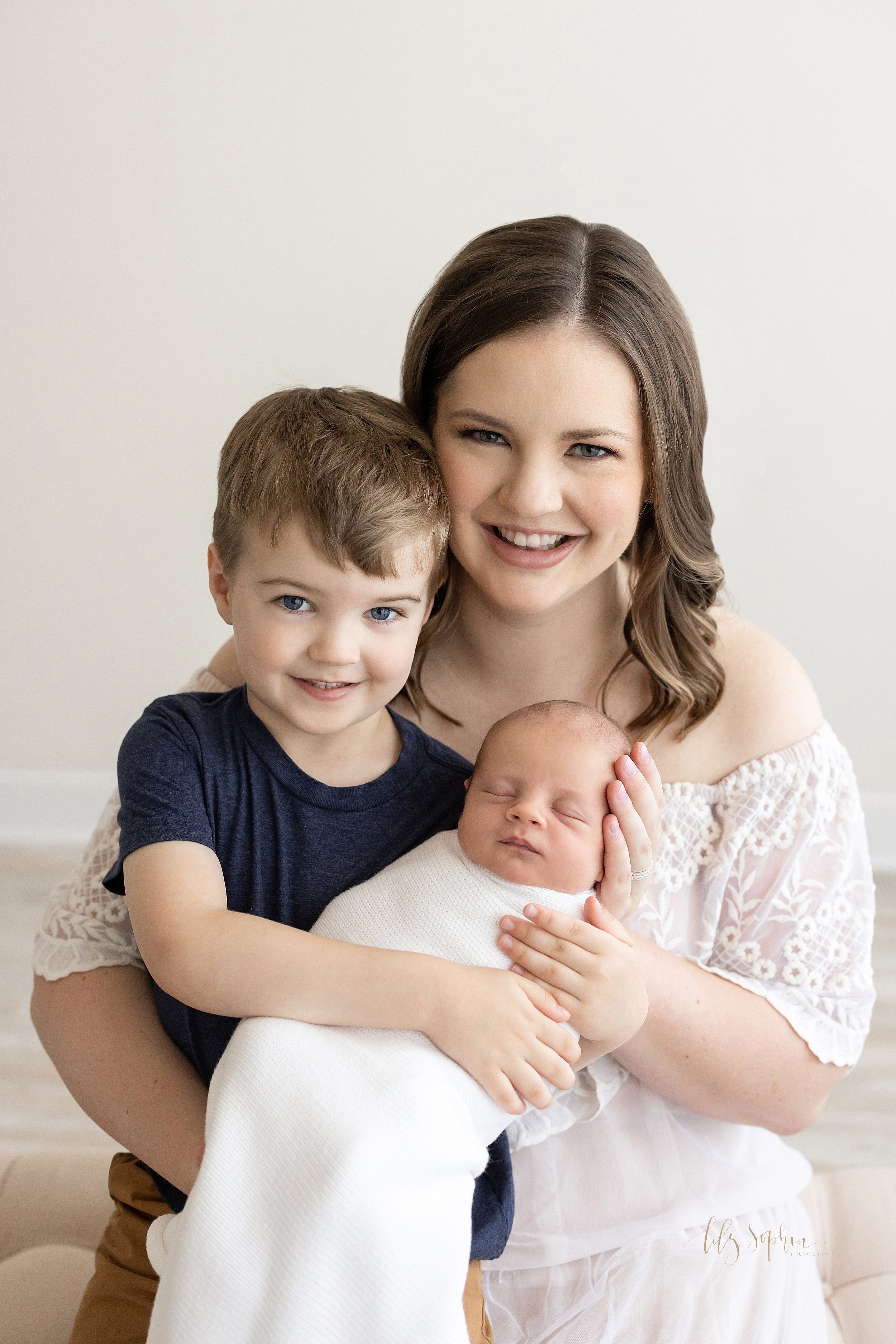  Family newborn portrait with mom sitting on an ottoman with her young son on her lap as the two of them hold the sleeping newborn baby boy taken in natural light near Smyrna in Atlanta in a photography studio. 