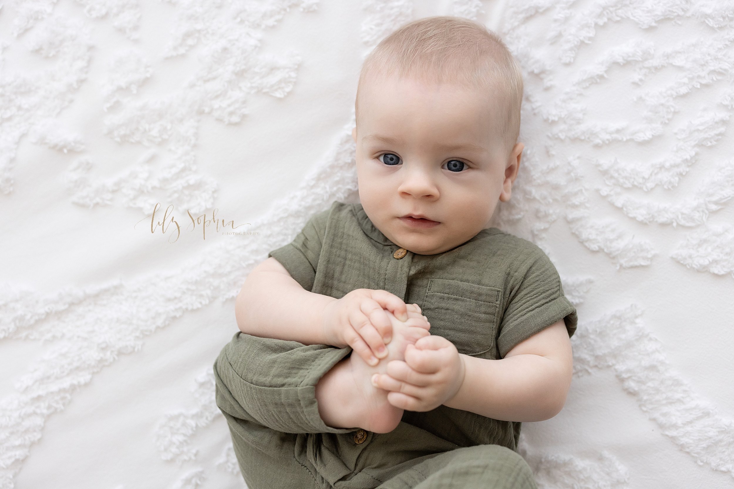  Overhead baby photo of a baby boy lying on his back on a bed and playing with his right foot taken using natural light in a studio near Poncey Highlands near Atlanta, Georgia.  