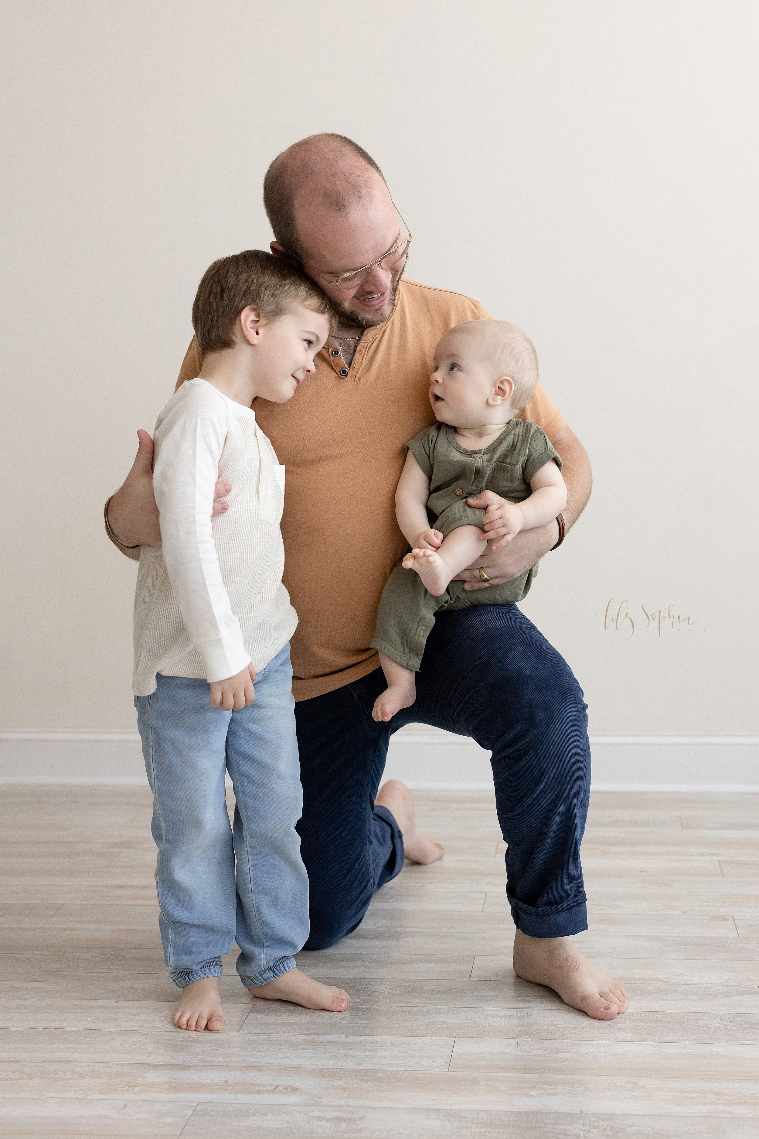  Family photo of a father kneeling on his right knee with his baby son sitting on his left knee as he looks up at his father while his brother stands in front of his father with his father’s hand around him as he looks at his baby brother taken in fr