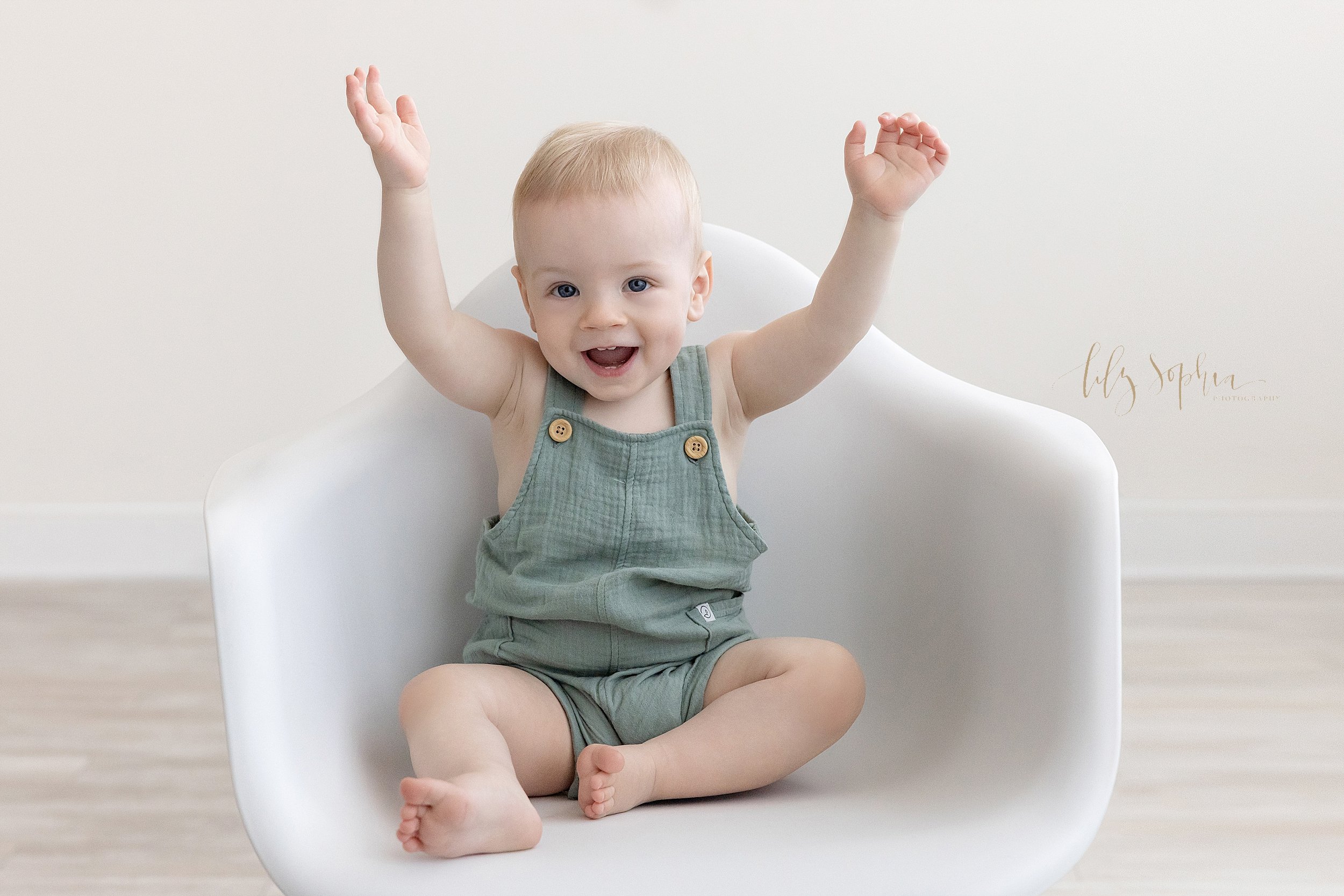  First birthday portrait of a one year old boy as he sits in a white molded chair and raises his hands saying, “SO BIG” in a studio in front of a window streaming natural light near Ansley Park in Atlanta, Georgia. 