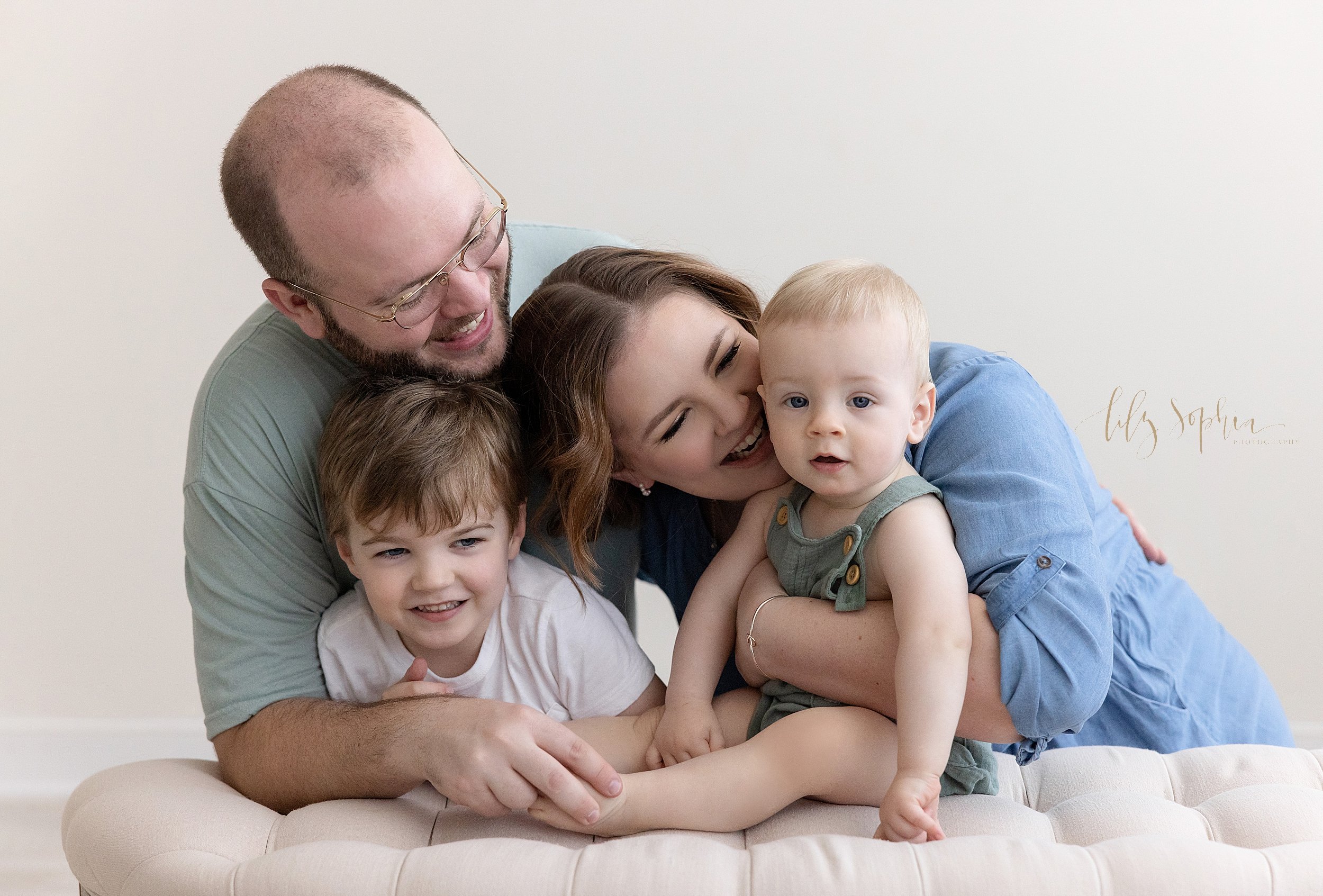  First birthday family picture of the one-year old boy sitting on top of a tufted ottoman with his mom holding him from behind as his brother kneels with his dad behind the ottoman and the family interacts taken in a studio near Buckhead in Atlanta t