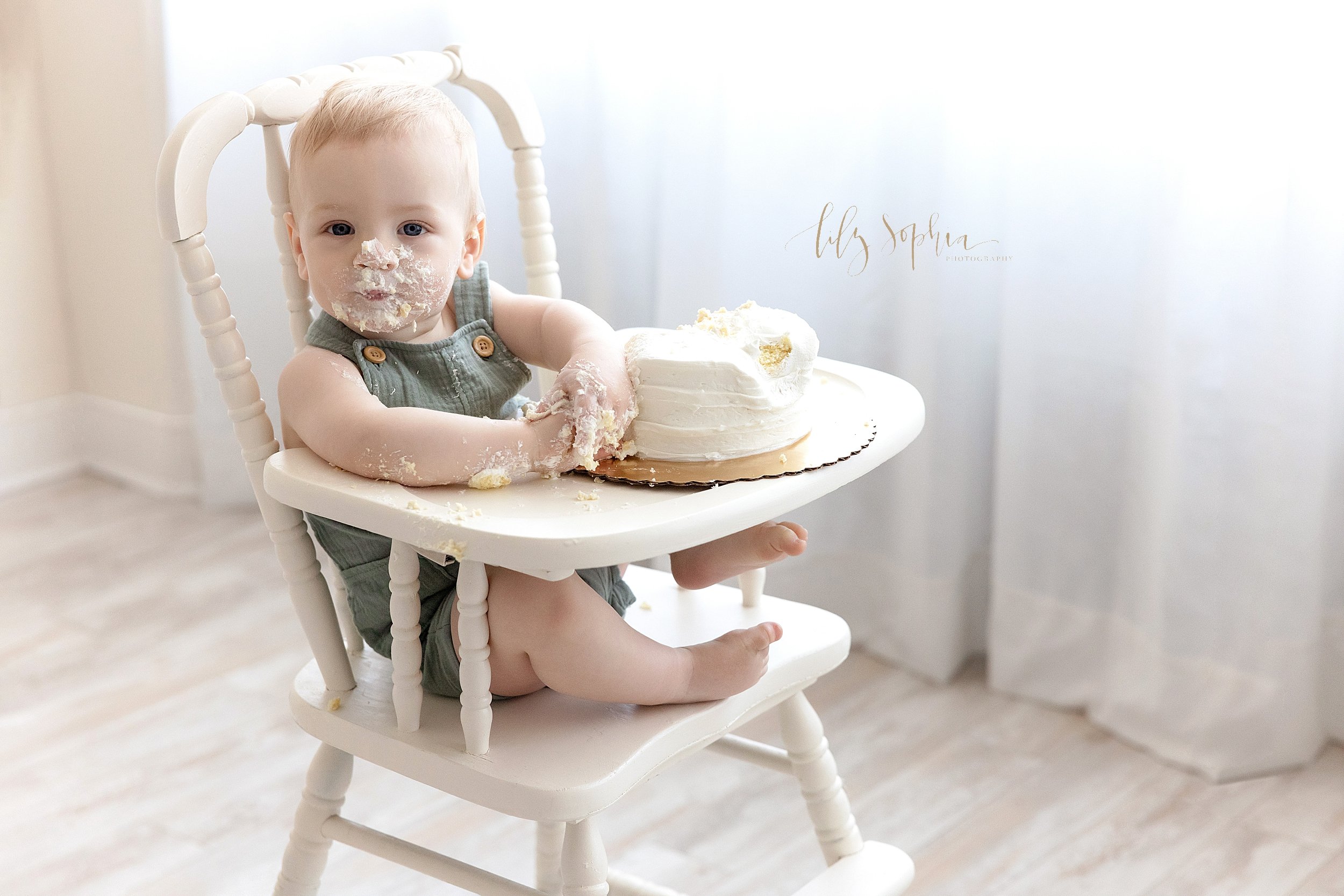  First birthday smash cake photo of a one year old little boy sitting in short overalls in an antique highchair with icing covering his hands and his face taken in front of a window streaming natural light in a studio near Sandy Springs in Atlanta, G