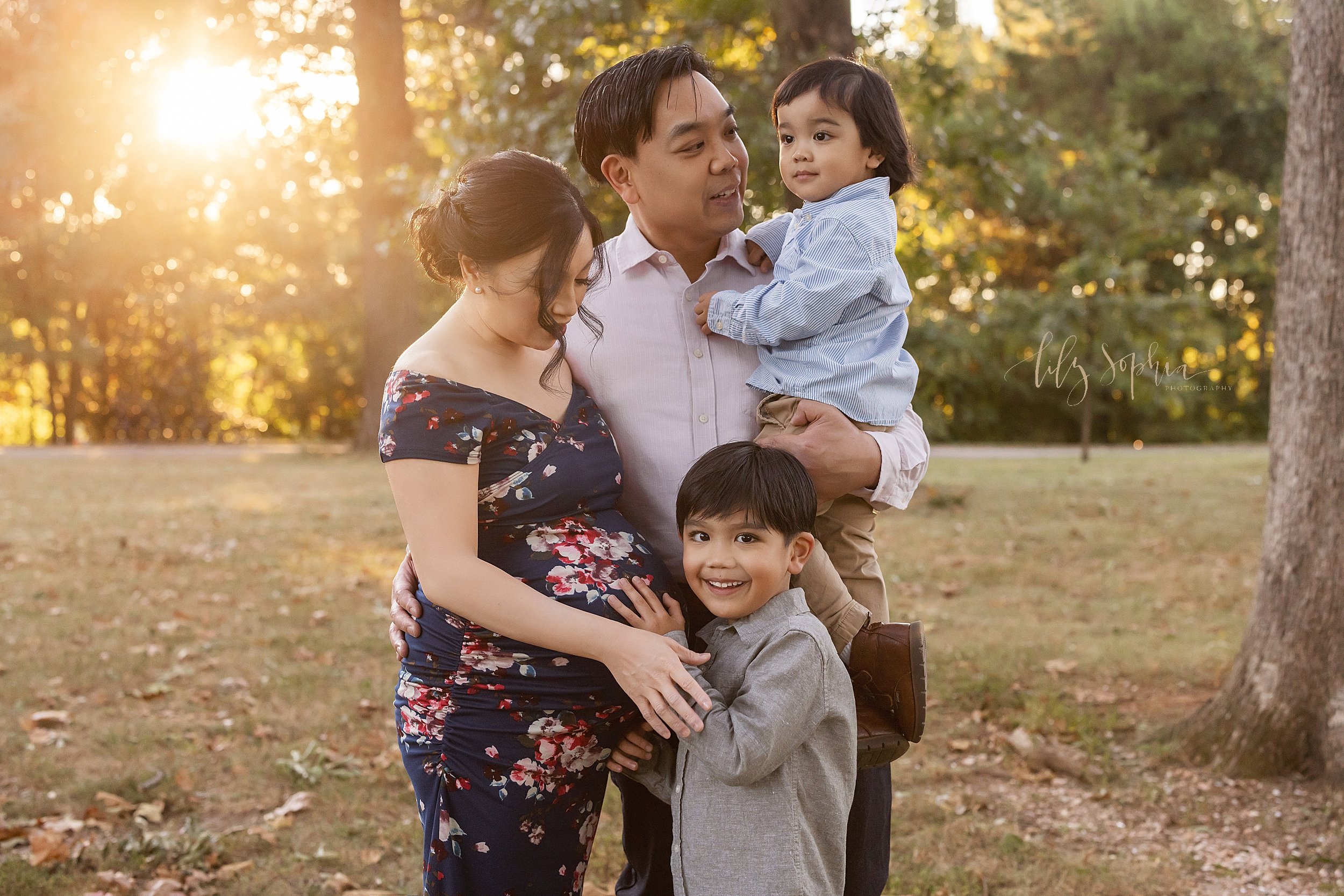  Maternity photo shoot of a mother wearing a floral knit gown with her husband standing to her left side and wrapping his right arm around her waist as he holds their young daughter in his left arm and their son places his right hand on his sibling i