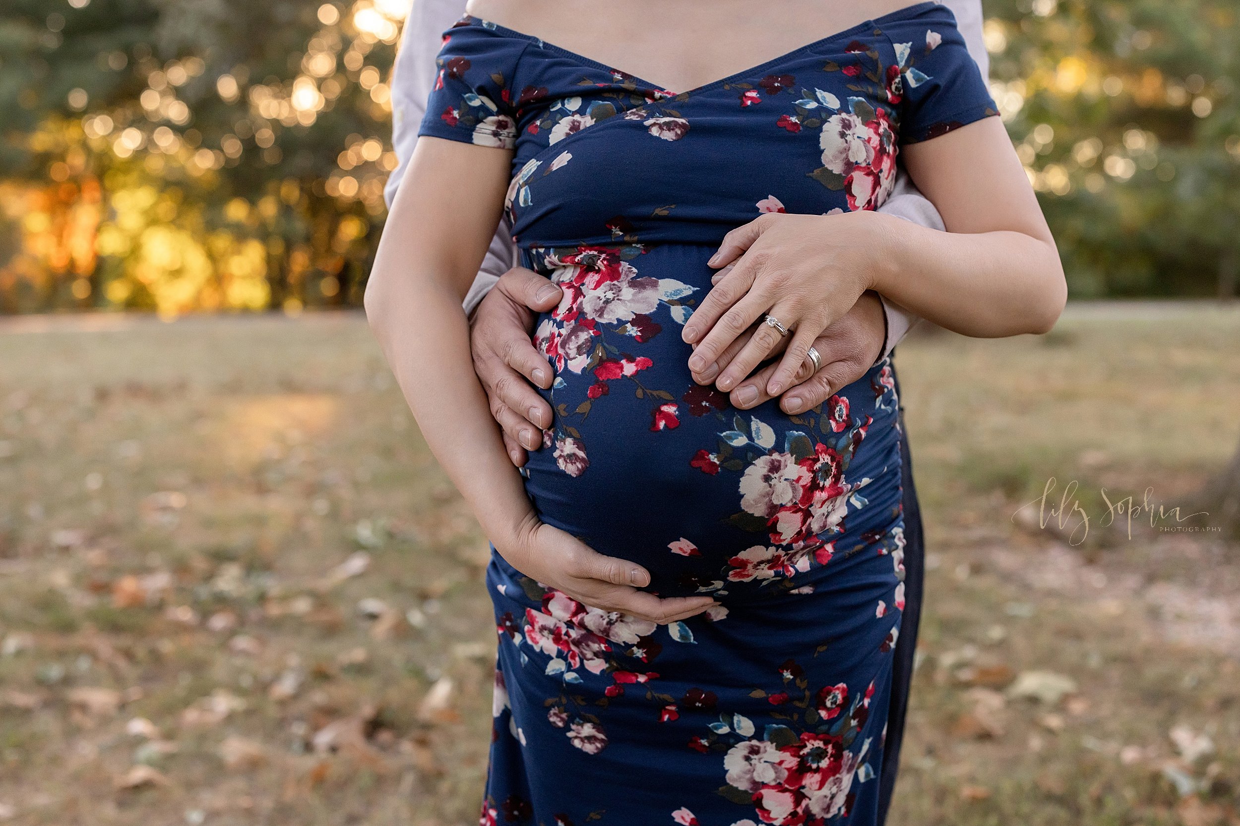  Maternity portrait of a pregnant woman’s belly with her husband standing behind her and placing his hands on their child in utero as the mother holds the base of her belly with her left hand and places her right hand over her husband’s taken at suns