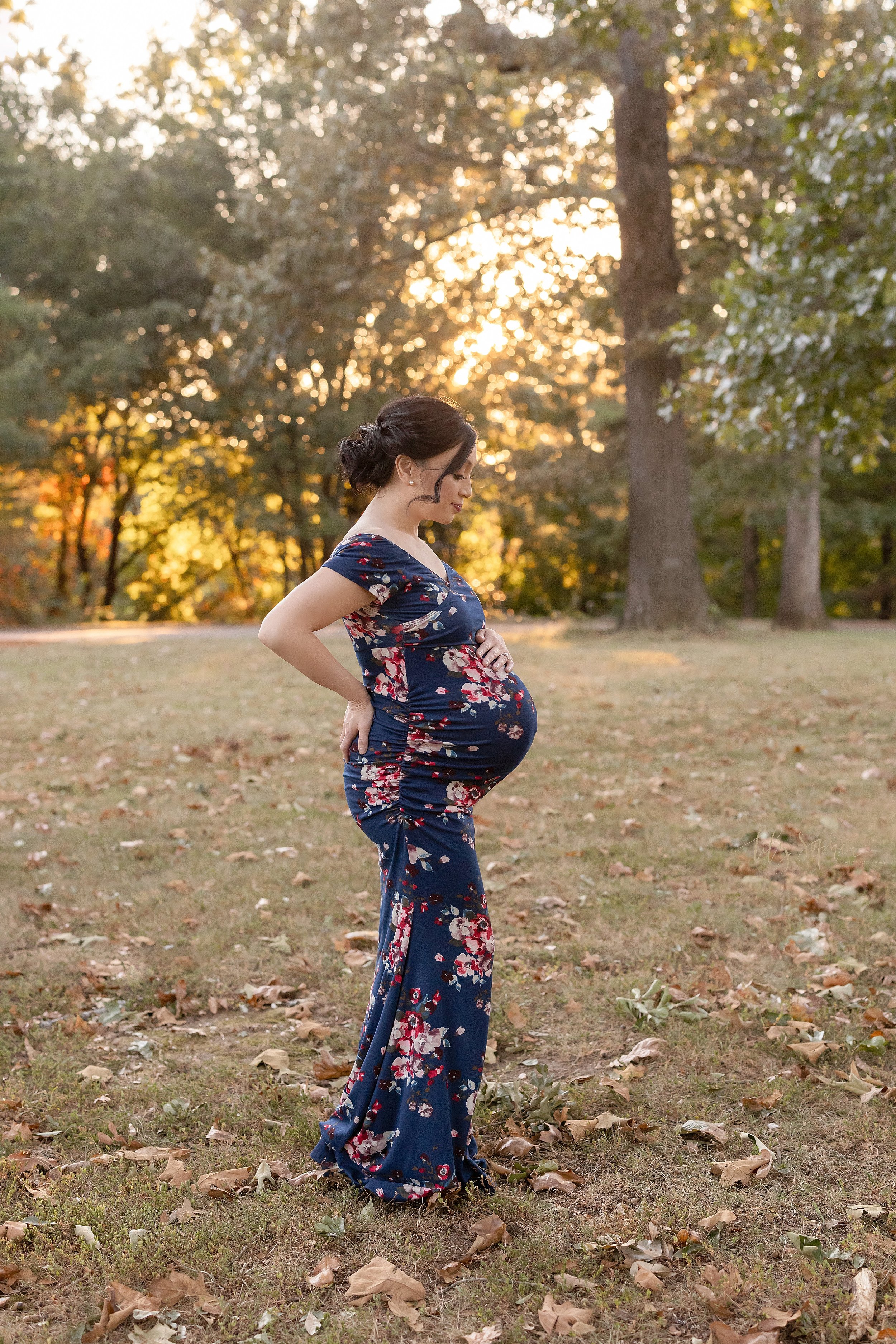  Maternity photo shoot of an Asian mother standing with her right hand on her hip and her left hand on the top of her belly as the sun sets behind her in an Atlanta park. 