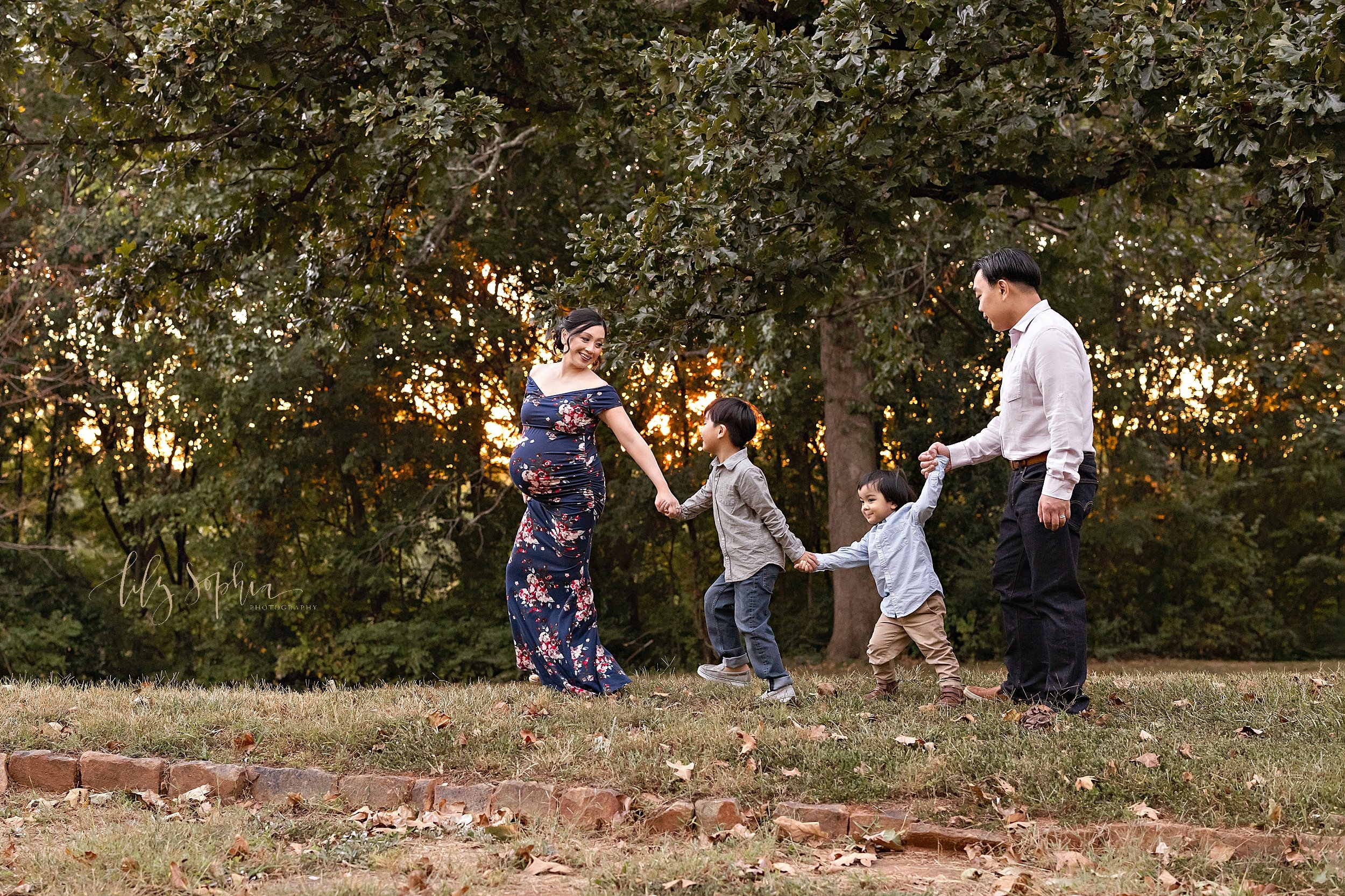  Family picture of a pregnant mother leading the way as she holds her son’s hand who holds his brother’s hand who holds his father’s hand as they walk through an Atlanta park playing follow the leader at sunset. 