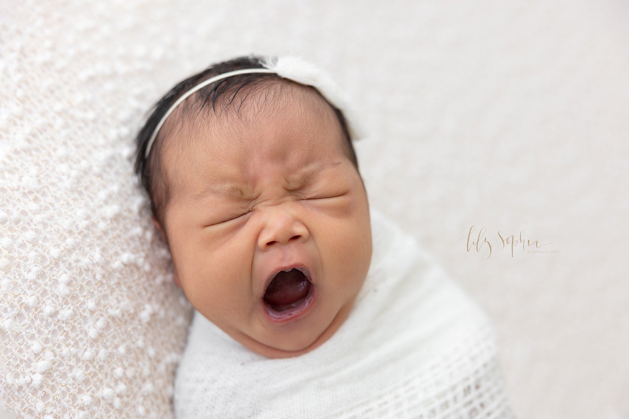  Newborn portrait of an Asian baby girl swaddled in a white knitted blanket and wearing a delicate headband in her hair as she opens her mouth to yawn taken in a natural light studio near Ponce City Market in Atlanta. 
