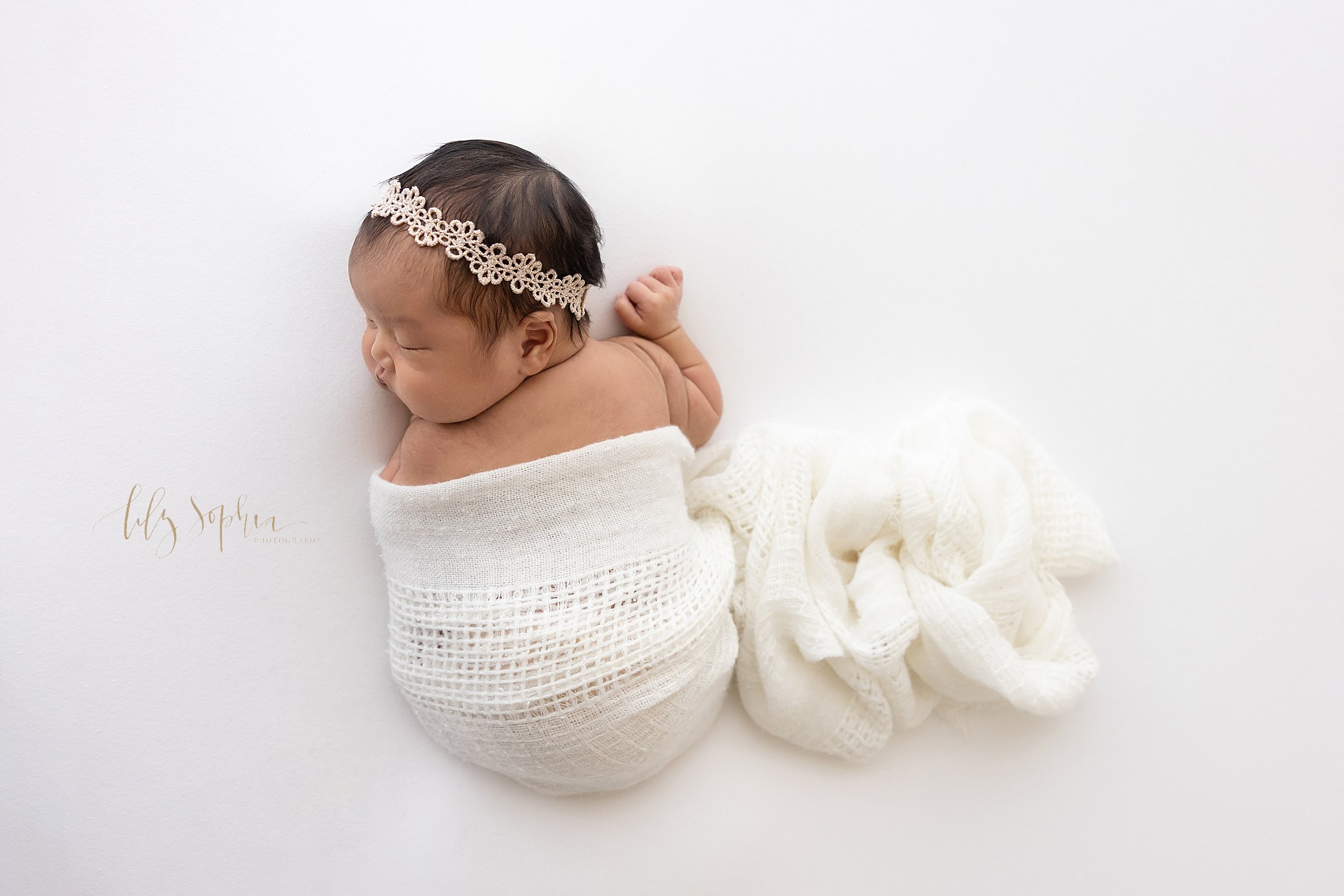  Newborn photo of a baby girl draped in a soft white knitted blanket lying on her stomach with her head turned to her right and wearing a delicate laurel headband on her head taken using natural light in a studio near Roswell in Atlanta. 