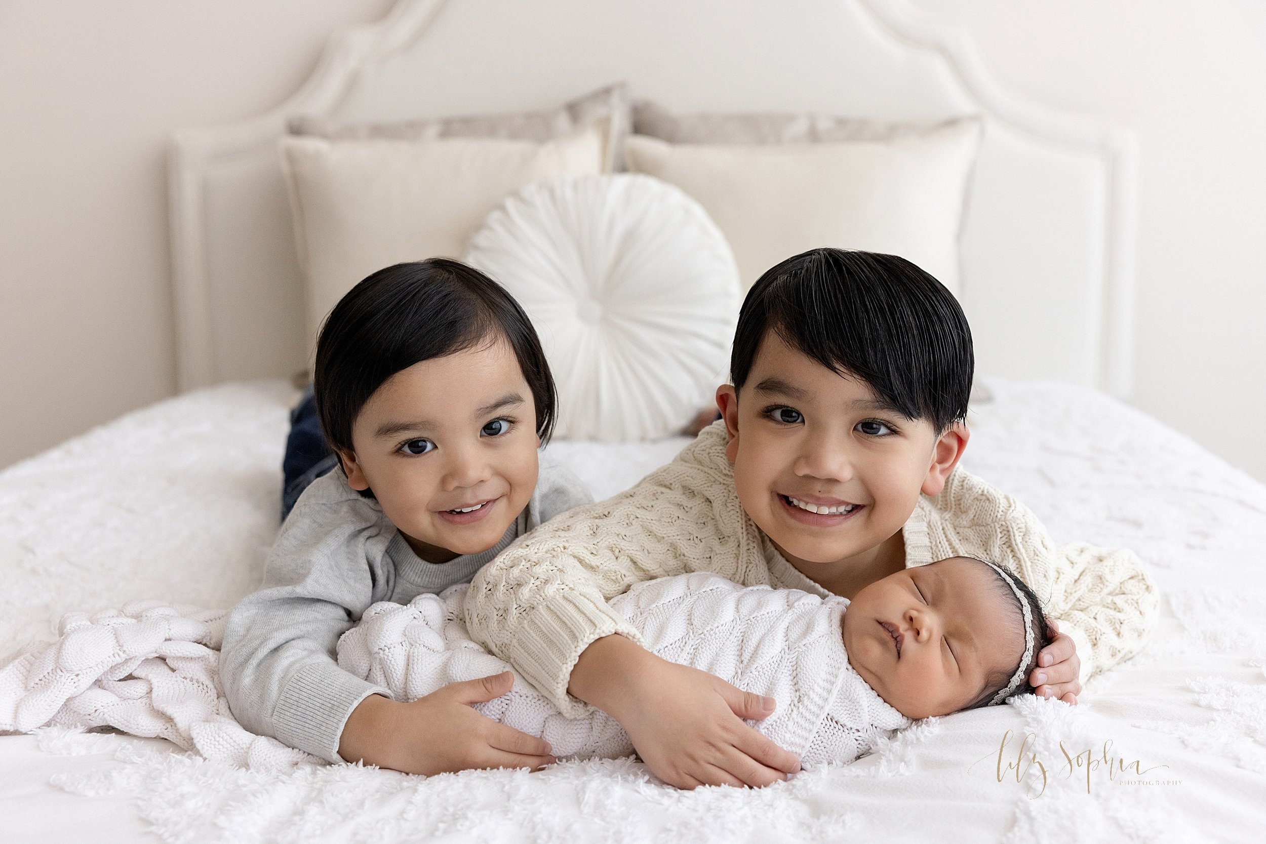  Newborn photo of Asian siblings lying on their stomachs on a bed while the older brother and sister hold their sleeping newborn baby sister taken in natural light in a studio near Midtown in Atlanta. 