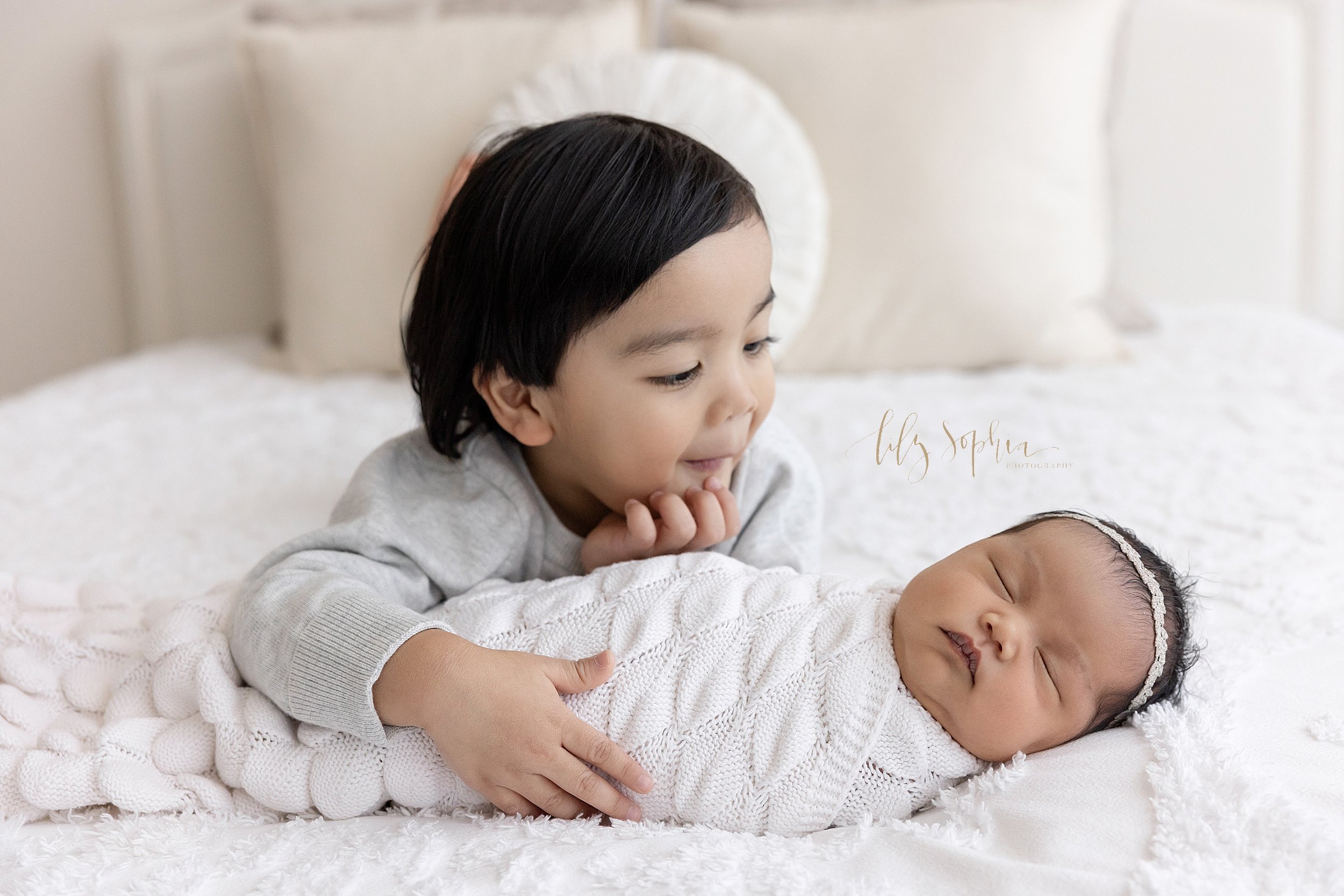  Newborn portrait of  sleeping newborn baby girl with her sister lying on her stomach and holding her baby sister with her right hand and admiring her as they lie on a bed in a studio near Vinings near Atlanta that uses natural light. 