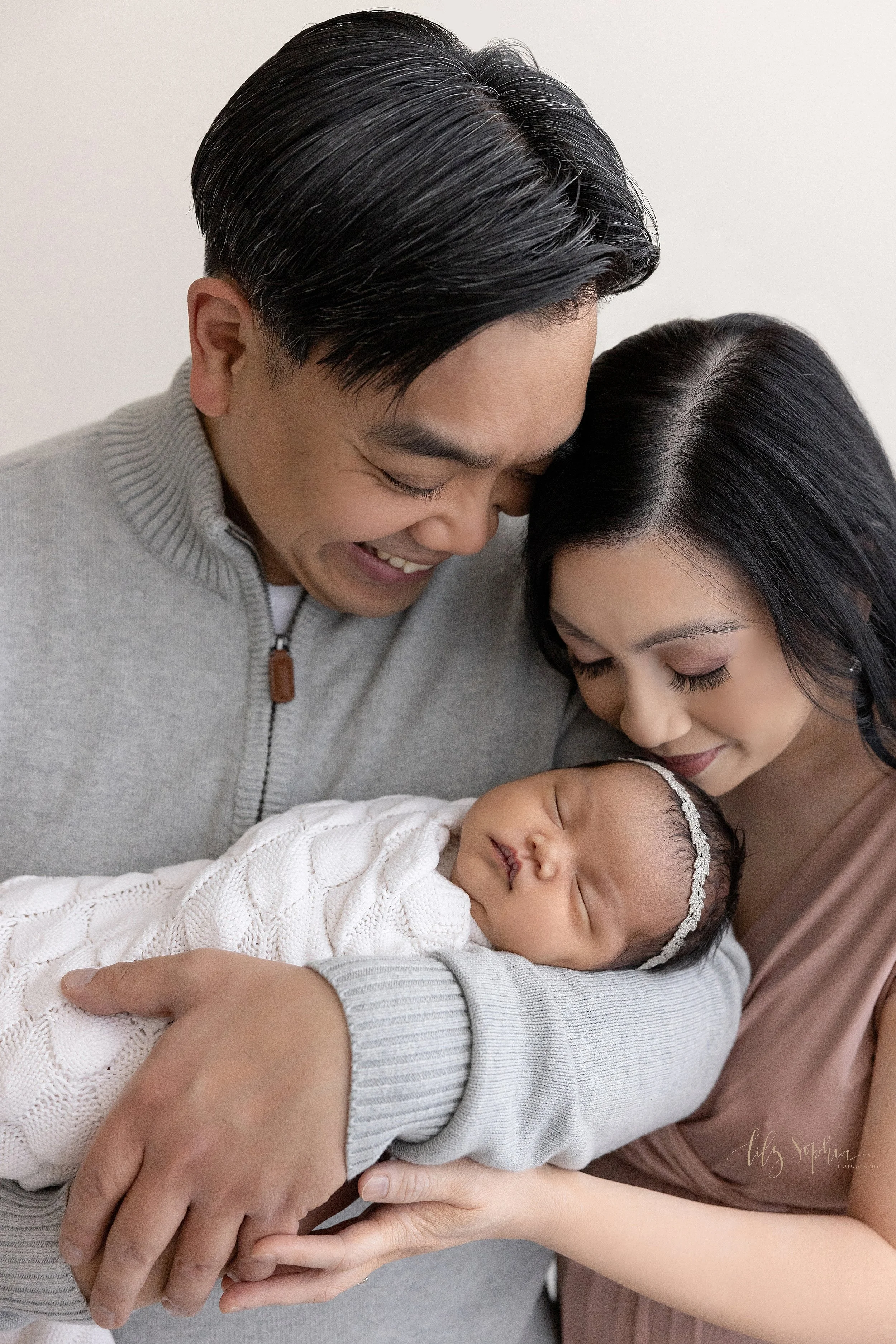  Newborn family portrait of an Asian father holding his newborn daughter in his arms as the mother stands to her husband’s right side and kisses her daughter’s head taken in a studio near Sandy Springs in Atlanta that uses natural light. 