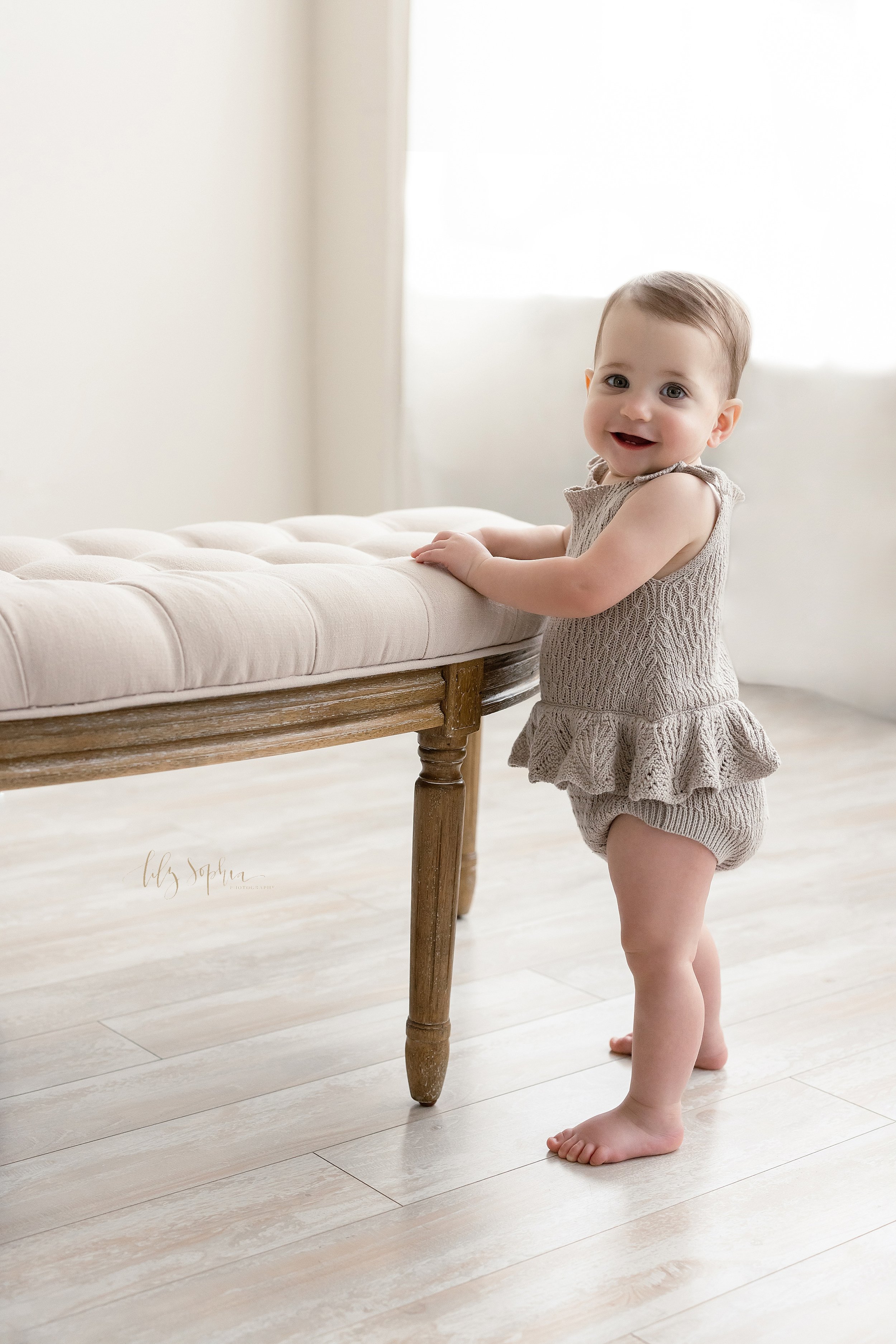  First birthday picture of a one year old baby girl standing while holding on to a tufted bench in front of a natural light window in a studio near Kirkwood in Atlanta, Georgia. 