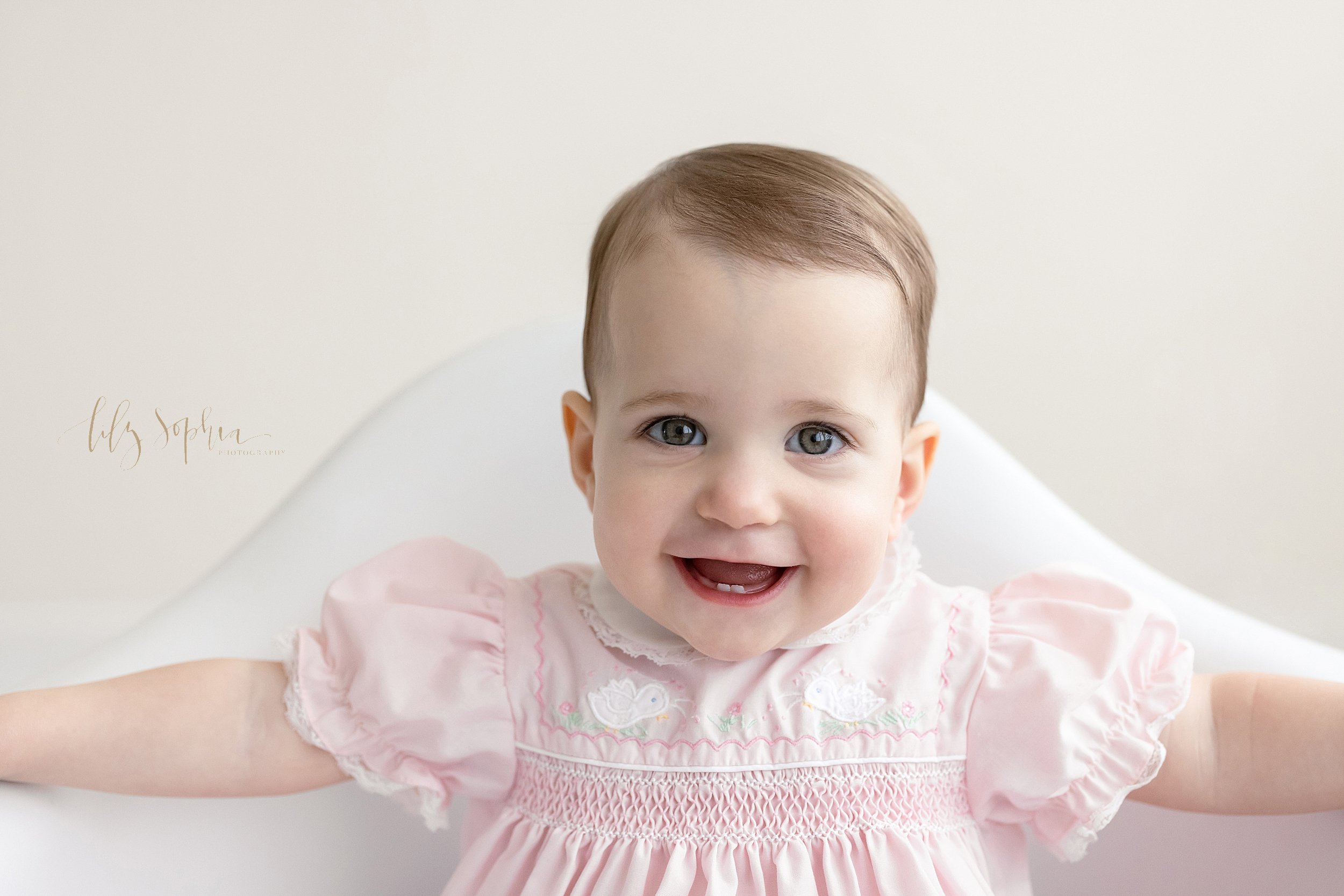  First Birthday close-up portrait of a smiling baby girl  wearing a smocked peter pan collared dress as she sits holding the sides of a white molded chair showing her baby teeth taken in a studio near Poncey Highlands in Atlanta that uses natural lig