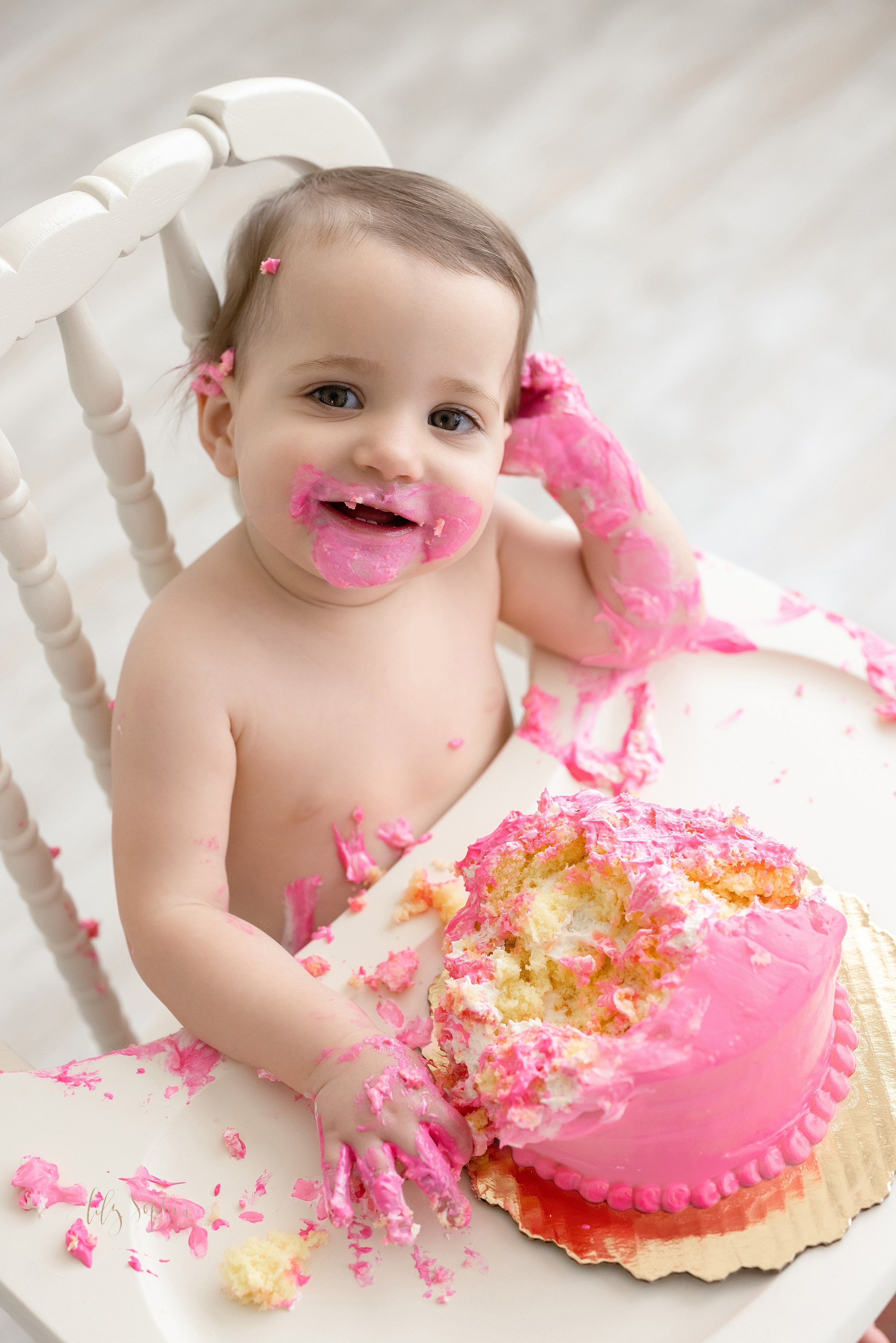  First birthday smash cake picture of a one year old little girl as she sits in an antique high chair smiling with pink icing surrounding her mouth and covering her right hand and arm taken in a natural light studio near Old Fourth Ward in Atlanta, G