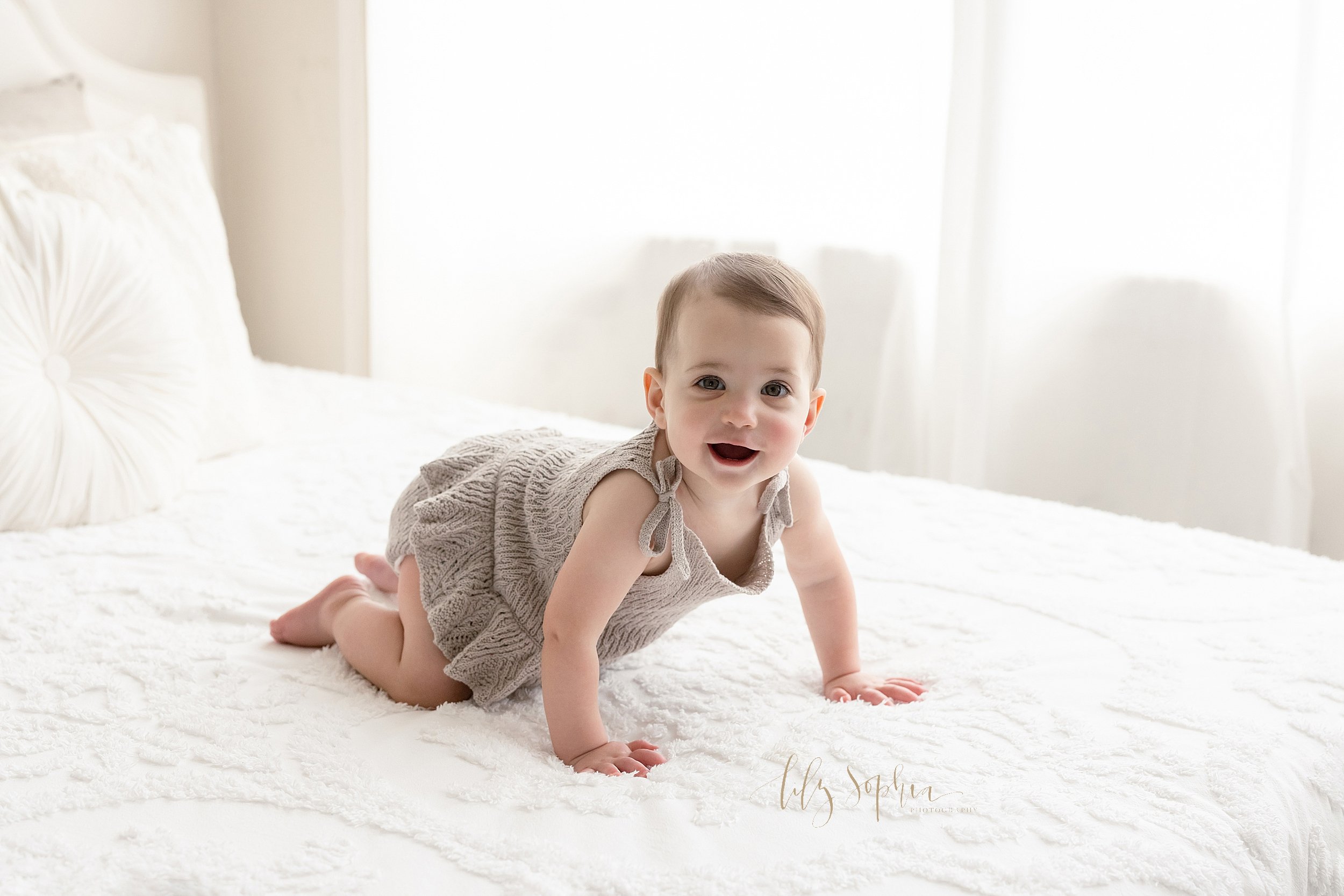  First birthday picture of a baby girl as she crawls in a knitted dress across a bed as natural light streams in from the window behind her in a studio near Smyrna in Atlanta. 