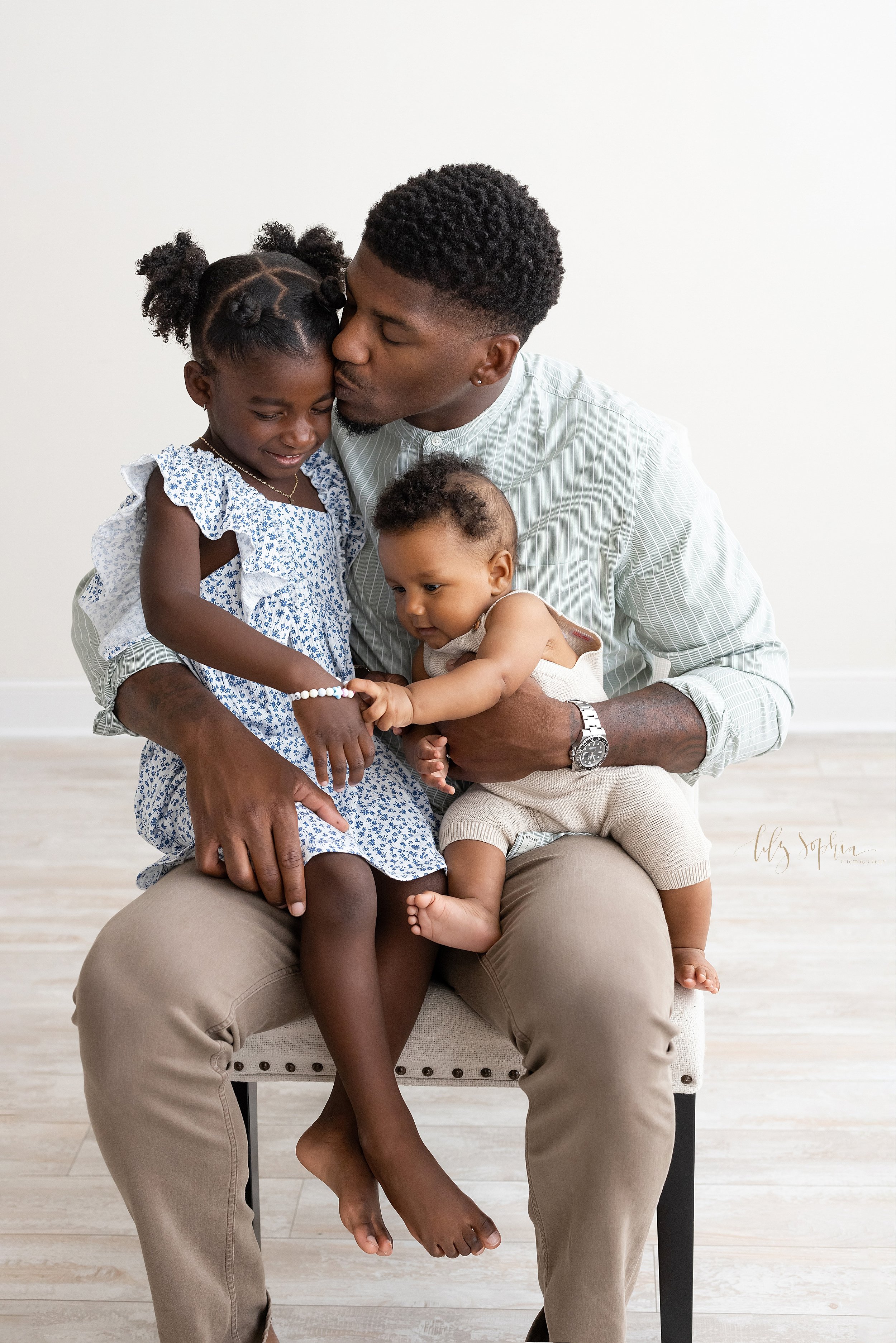  Family photo shoot of an African-American father sitting in an upholstered chair with his young daughter on his right knee and his baby son on his left knee taken using natural light in a studio near Old Fourth Ward in Atlanta, Georgia. 