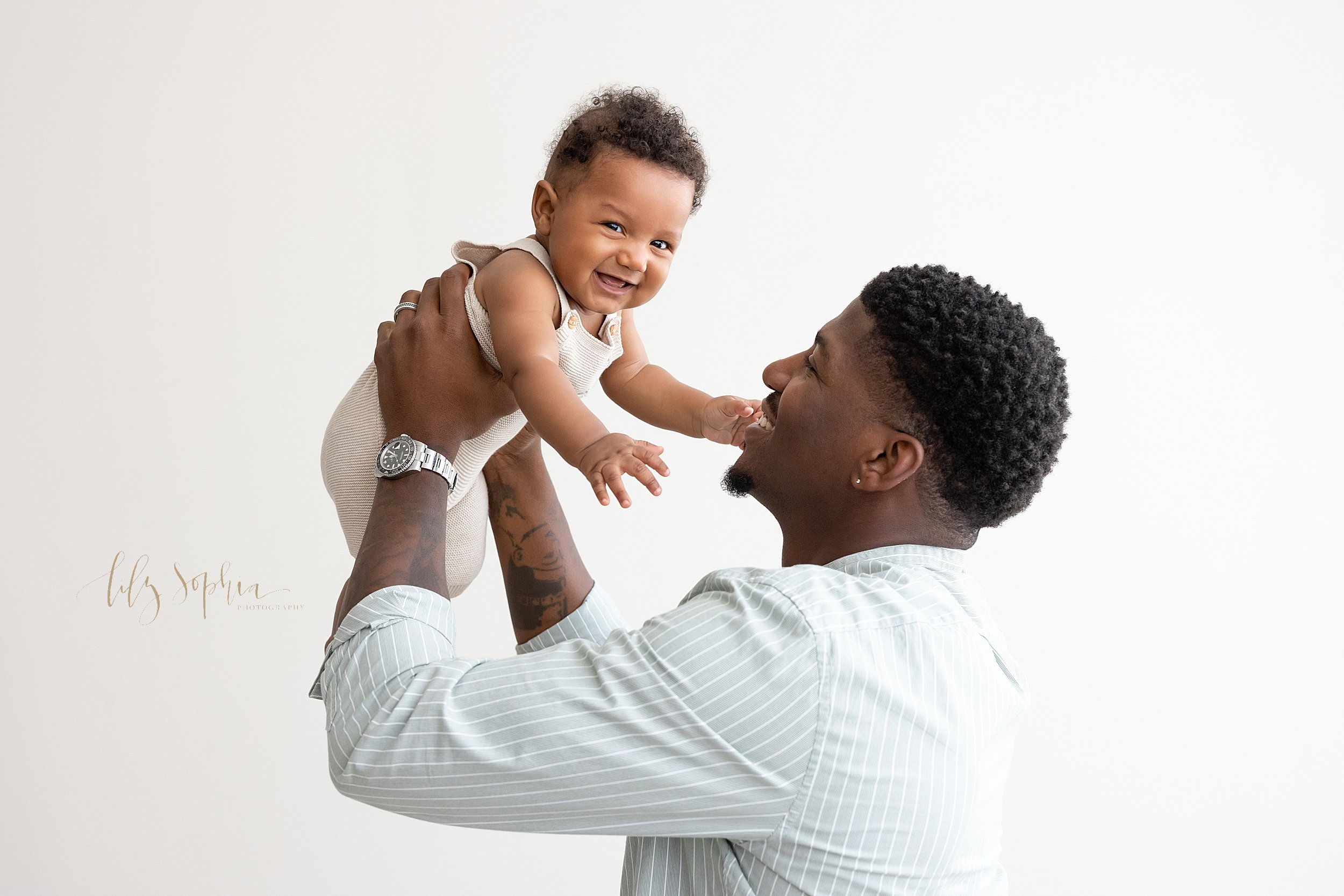  Family photo shoot of an African-American father holding his baby son in his hands above his head as he stands in front of a window streaming natural light in a studio near Oakhurst in Atlanta. 