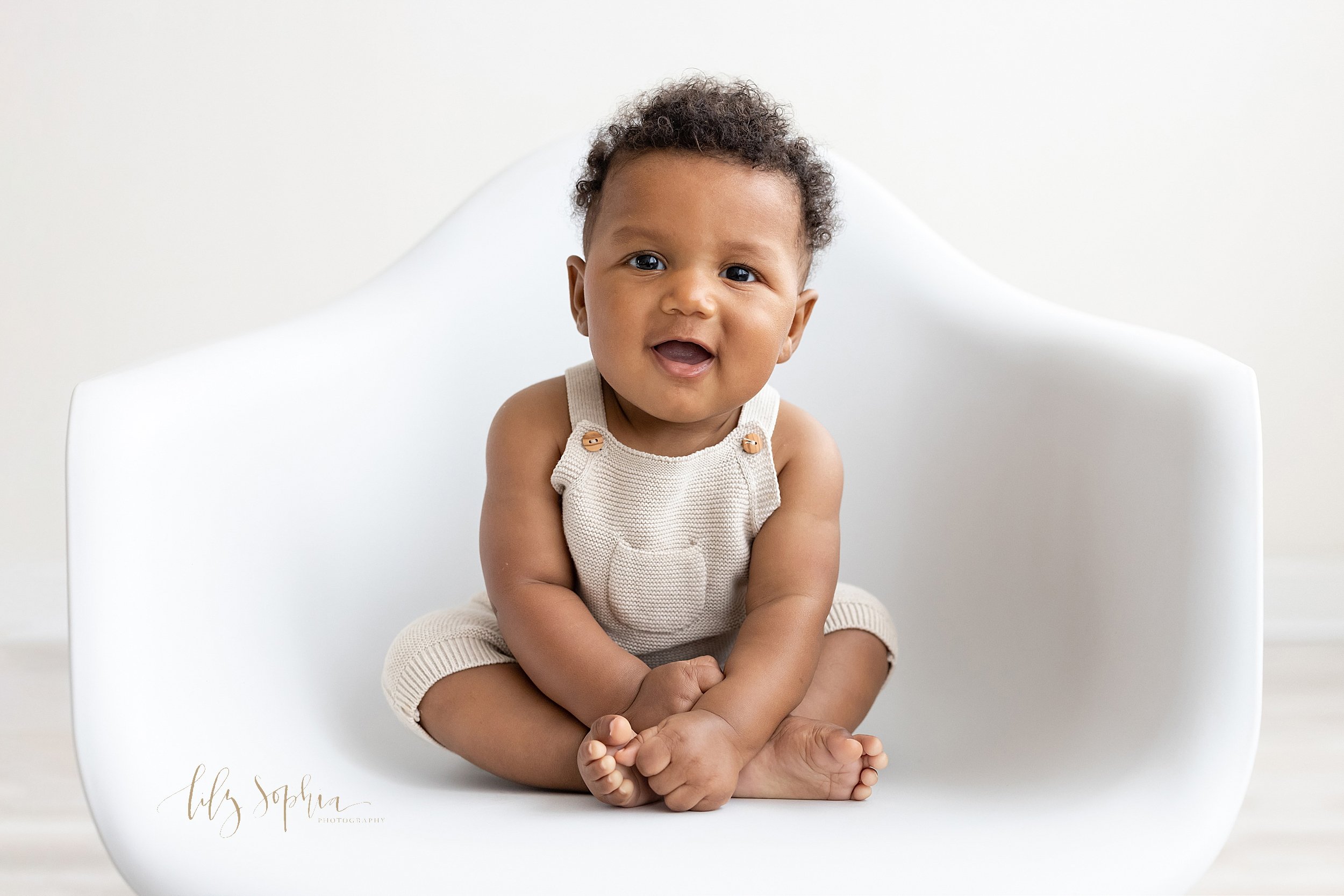  Baby portrait of an African-American boy as he sits cross-legged in a white molded chair holding his feet taken in a studio near Midtown in Atlanta that uses natural light. 