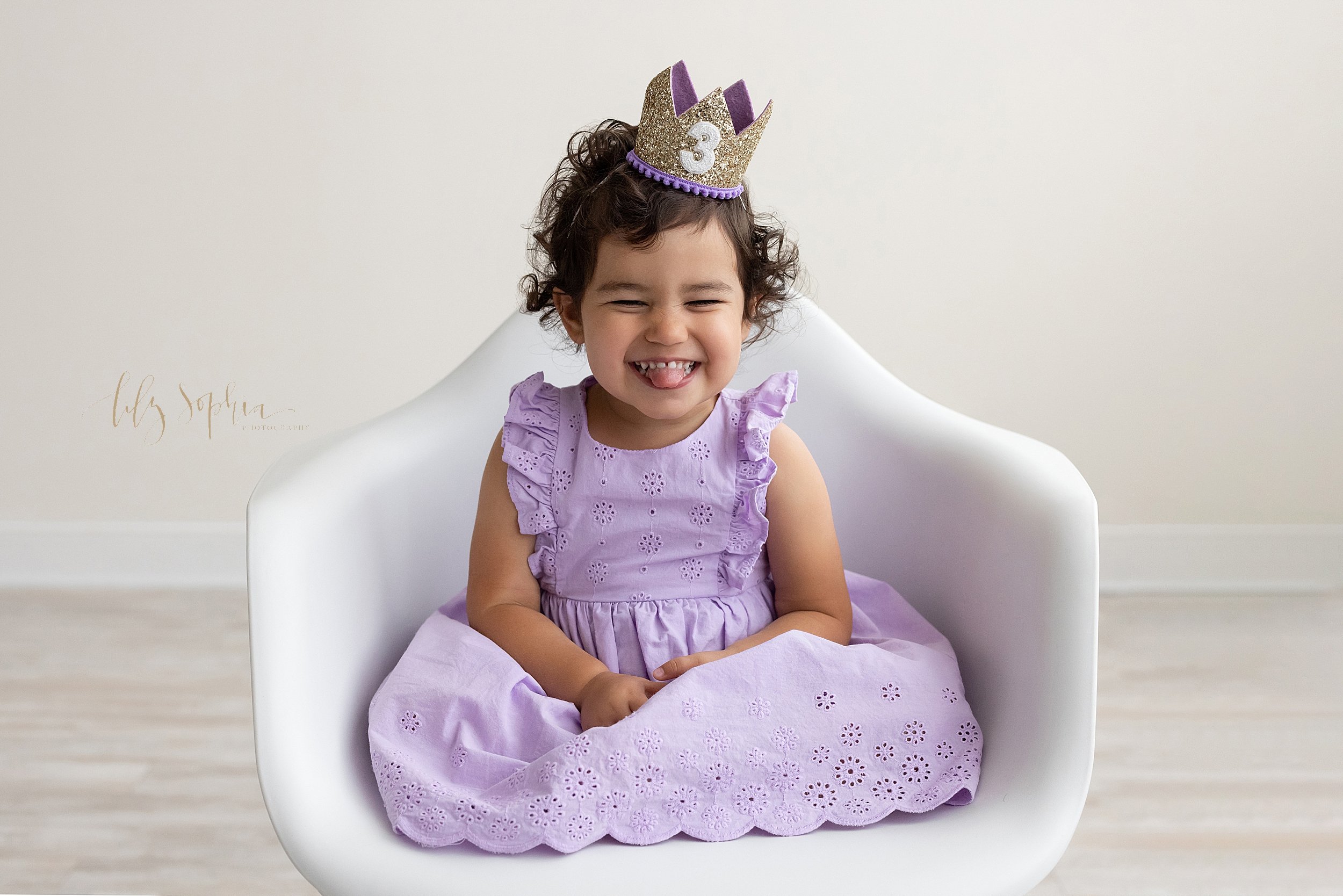  Birthday portrait of a three year old little girl sitting a  white molded chair wearing a lilac eyelet dress and a number 3 crown on her head as she sticks out her tongue while giggling taken in a natural light studio near Brookhaven in Atlanta, Geo