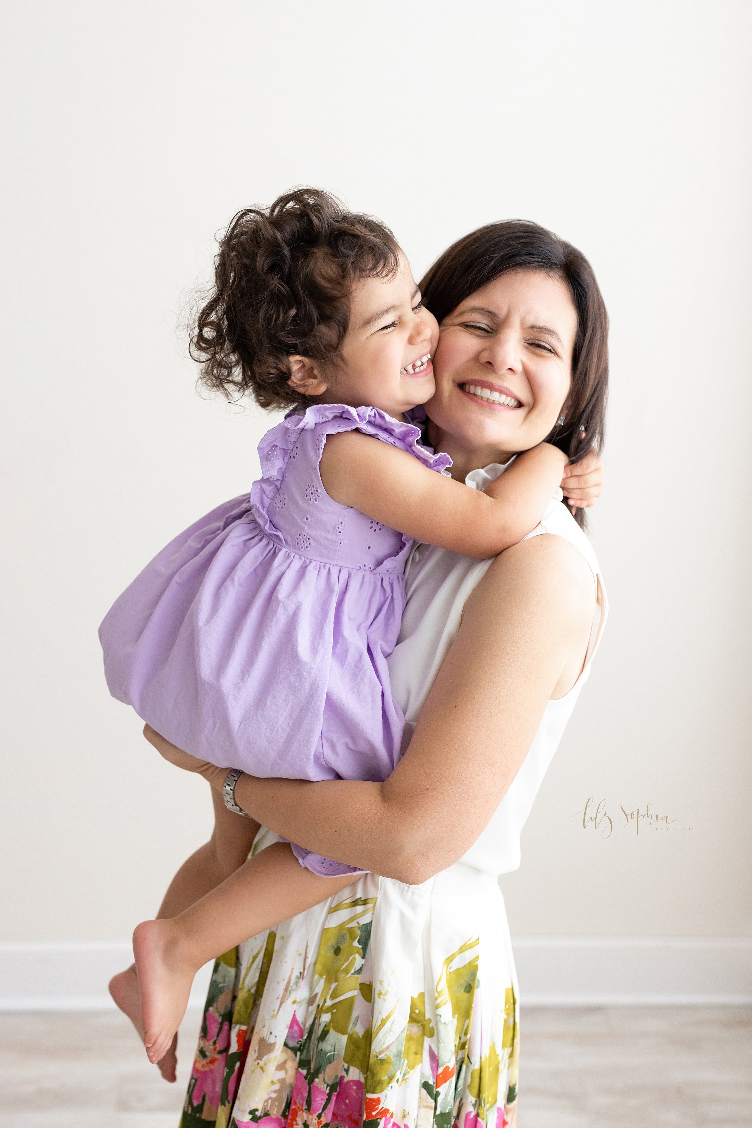  First birthday portrait of a happy three year old squeezing the neck of her mother as she holds her while standing in a natural light studio near Cummings in Atlanta. 