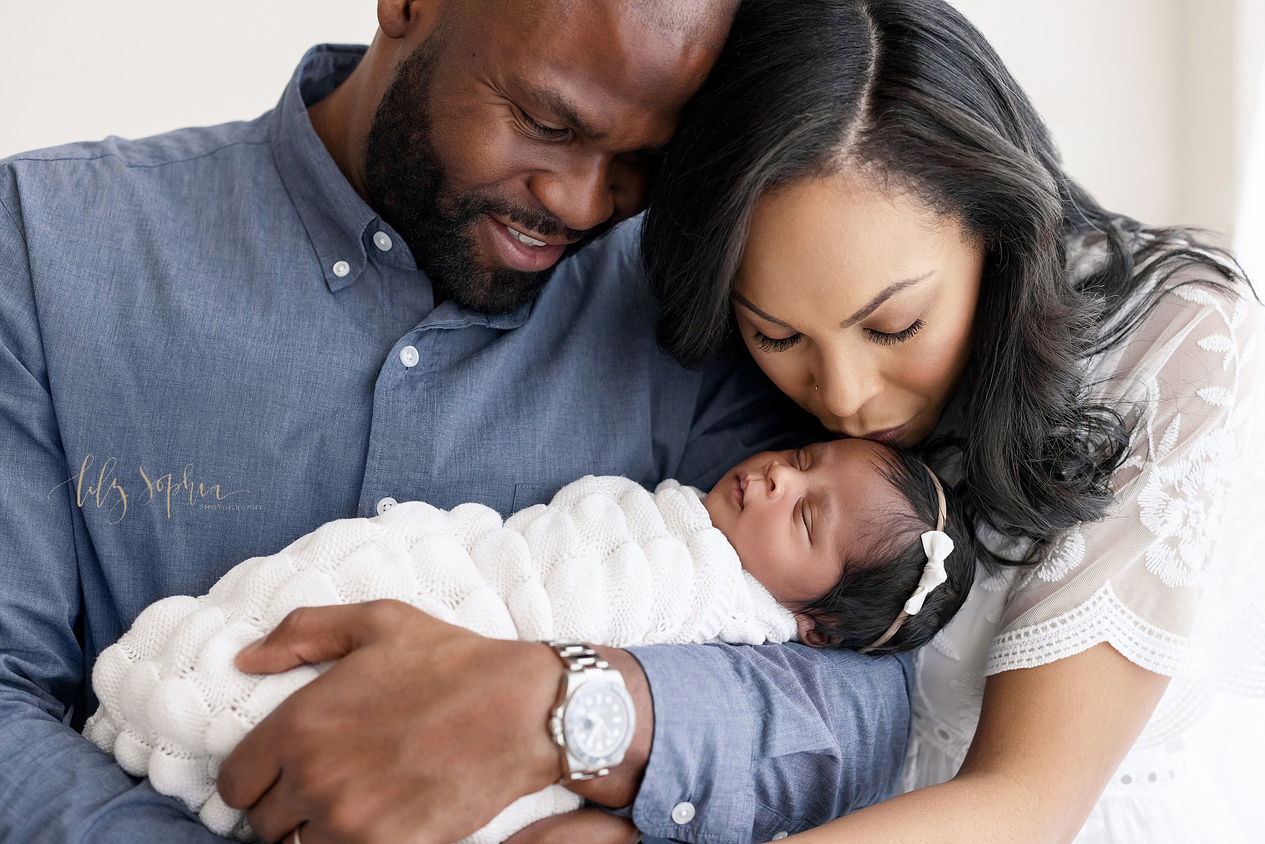  Newborn photo shoot of an African-American father holding his peacefully sleeping daughter in his arms in front of his chest while mom bends over and kisses her daughter’s forehead taken in natural light in a studio near Ansley Park near Atlanta, Ge