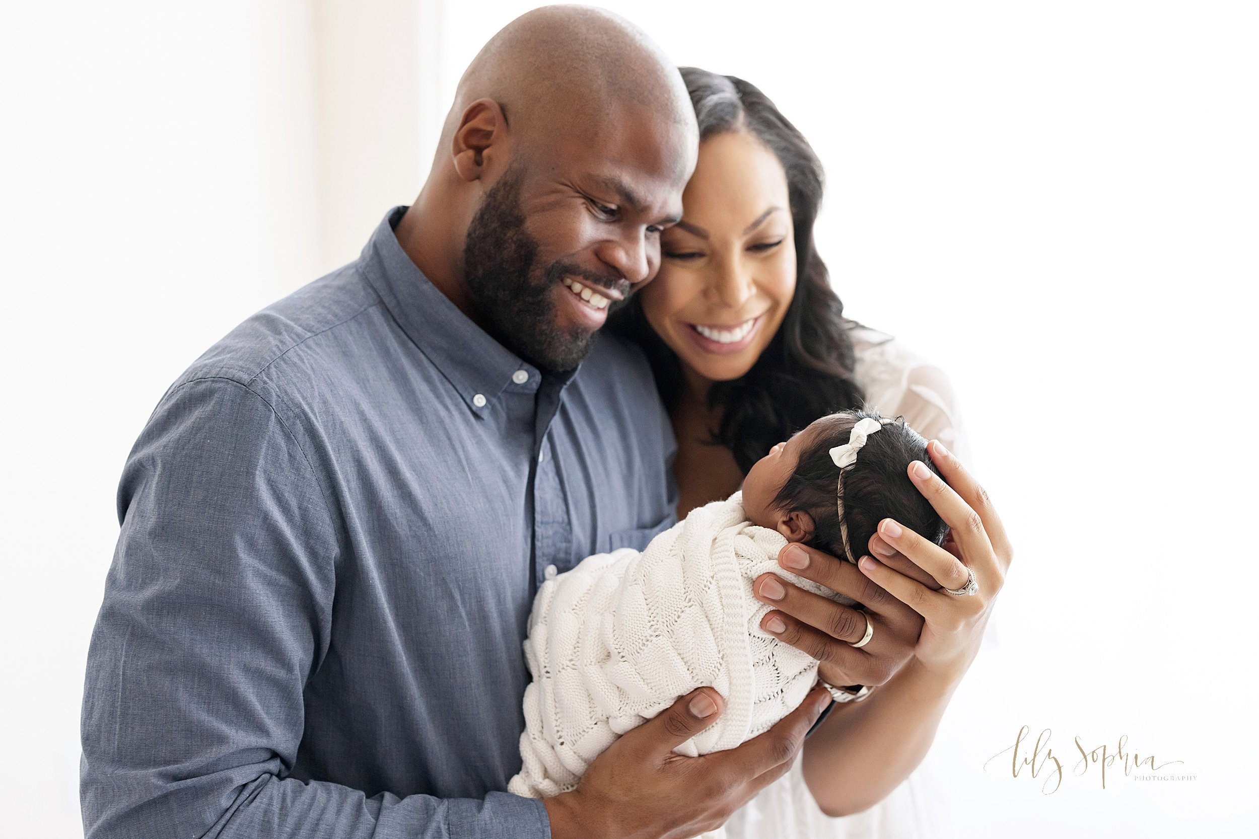  Newborn picture of an ecstatic African-American mother and father as dad stands holding his daughter’s head in his hand and mom stands next to him placing her hand on the crown of her daughter’s head in front of a window streaming natural light in a