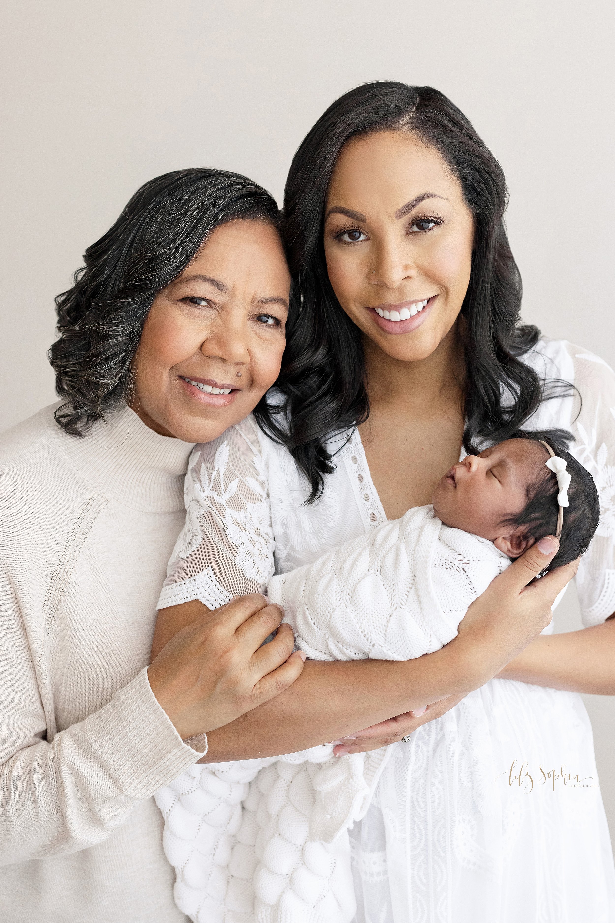  Intergenerational newborn photograph with the mother holding her infant daughter in her arms as the grandmother holds on to her daughter’s right arm while the two of them stand in a studio near Brookhaven in Atlanta that uses natural light. 