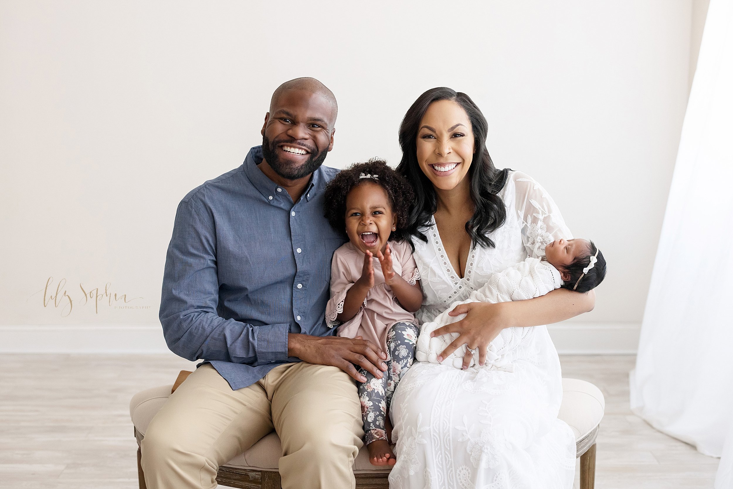  Newborn photo shoot with an African-American family sitting on a tufted ottoman with dad and mom sitting next to one another with their toddler daughter between them and mom holding their newborn baby girl in her left arm taken in a natural light st