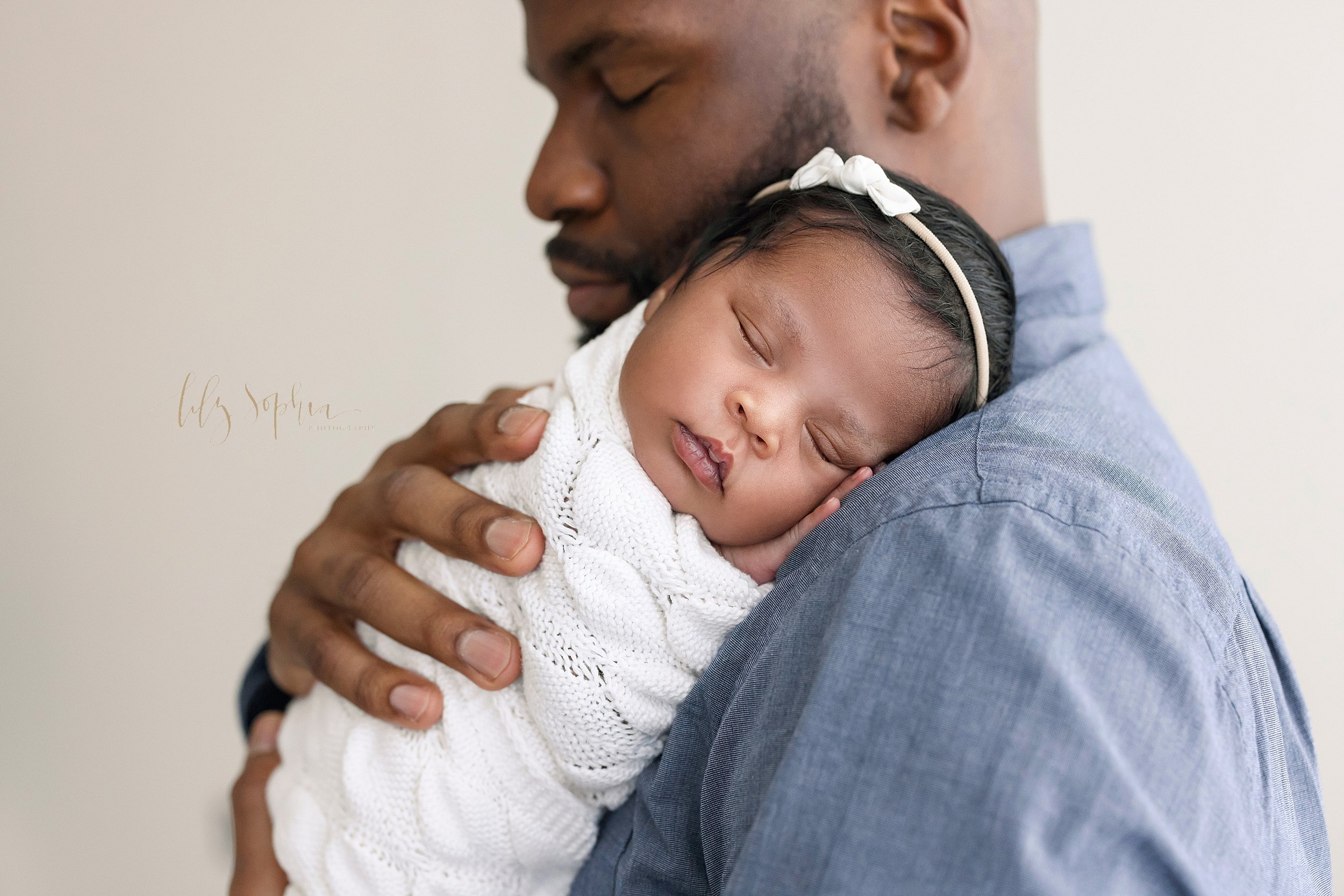  An African-American father holds his peacefully sleeping daughter wrapped in a soft white knitted blanket and wearing a delicate bow headband in her hair on his shoulder treasuring these special moments while standing in a studio near Decatur in Atl
