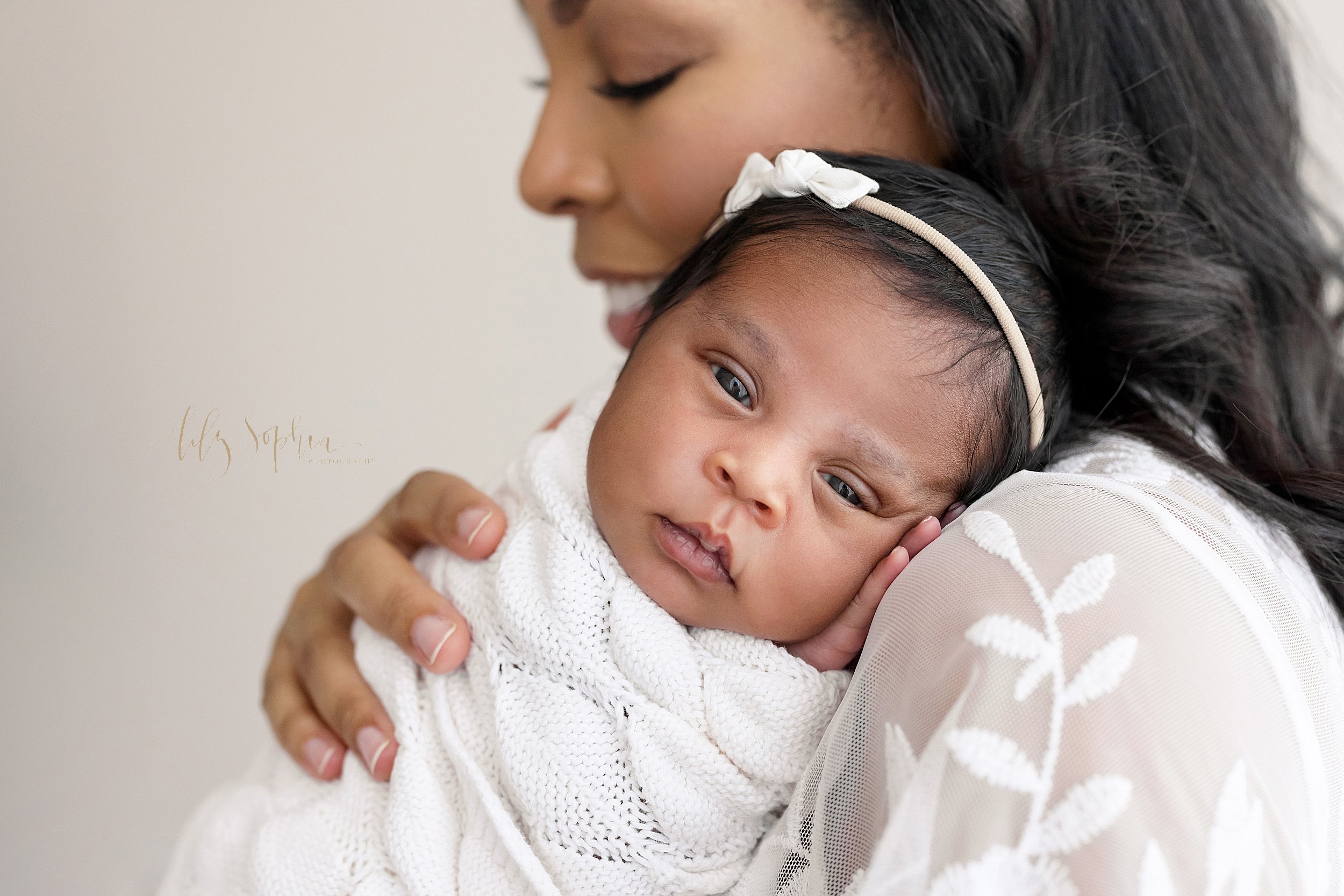  Newborn portrait of an African-American mother holding her awake infant baby girl wrapped in a soft white knitted blanket and wearing a delicate headband with a bow in her hair taken using natural light in a studio near Alpharetta in Atlanta. 