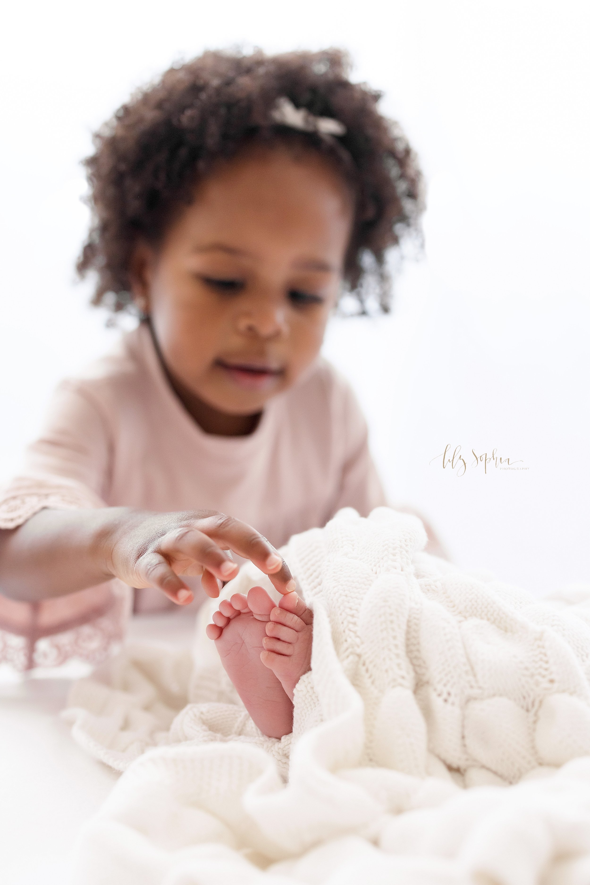  Newborn photo of a baby girl’s feet peeking out from a soft white knitted blanket as her toddler sister plays “This Little Piggy Goes to Market” on them taken in front of a window streaming natural light in a studio near Buckhead in Atlanta, Georgia