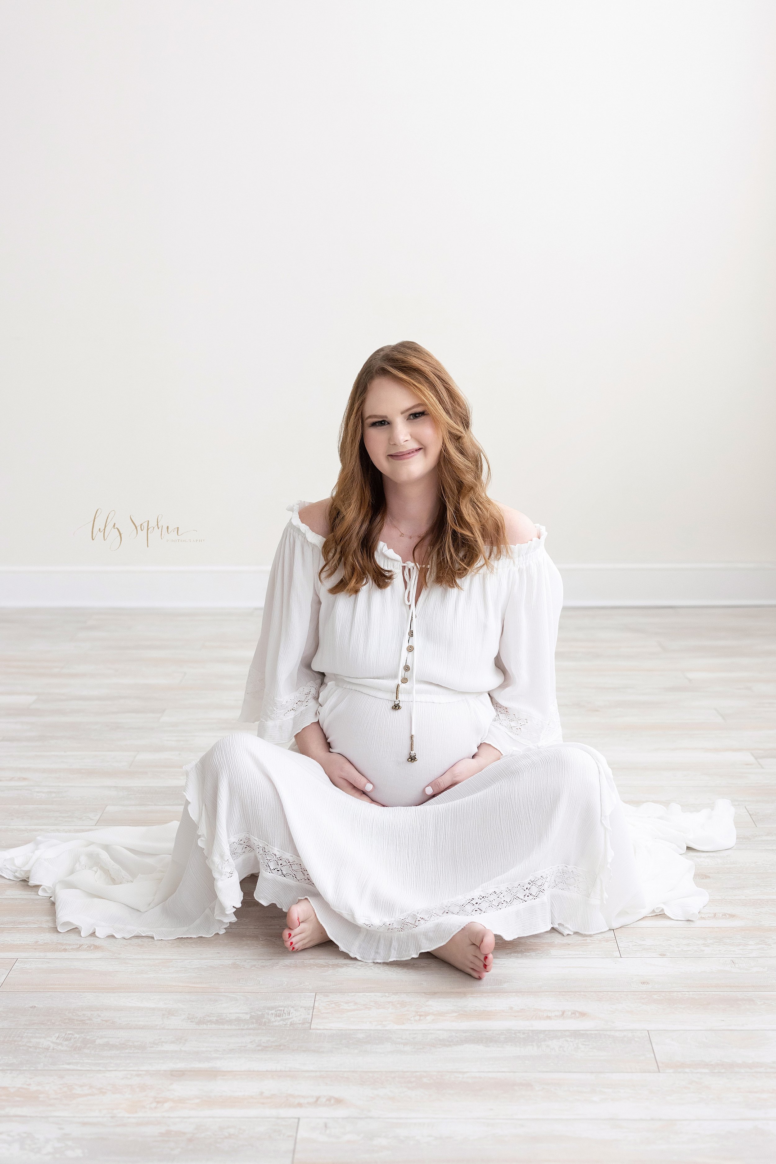 Maternity portrait of a pregnant mother sitting cross legged on the floor of a natural light studio with her ands holding her belly taken near Poncey Highlands in Atlanta, Georgia. 