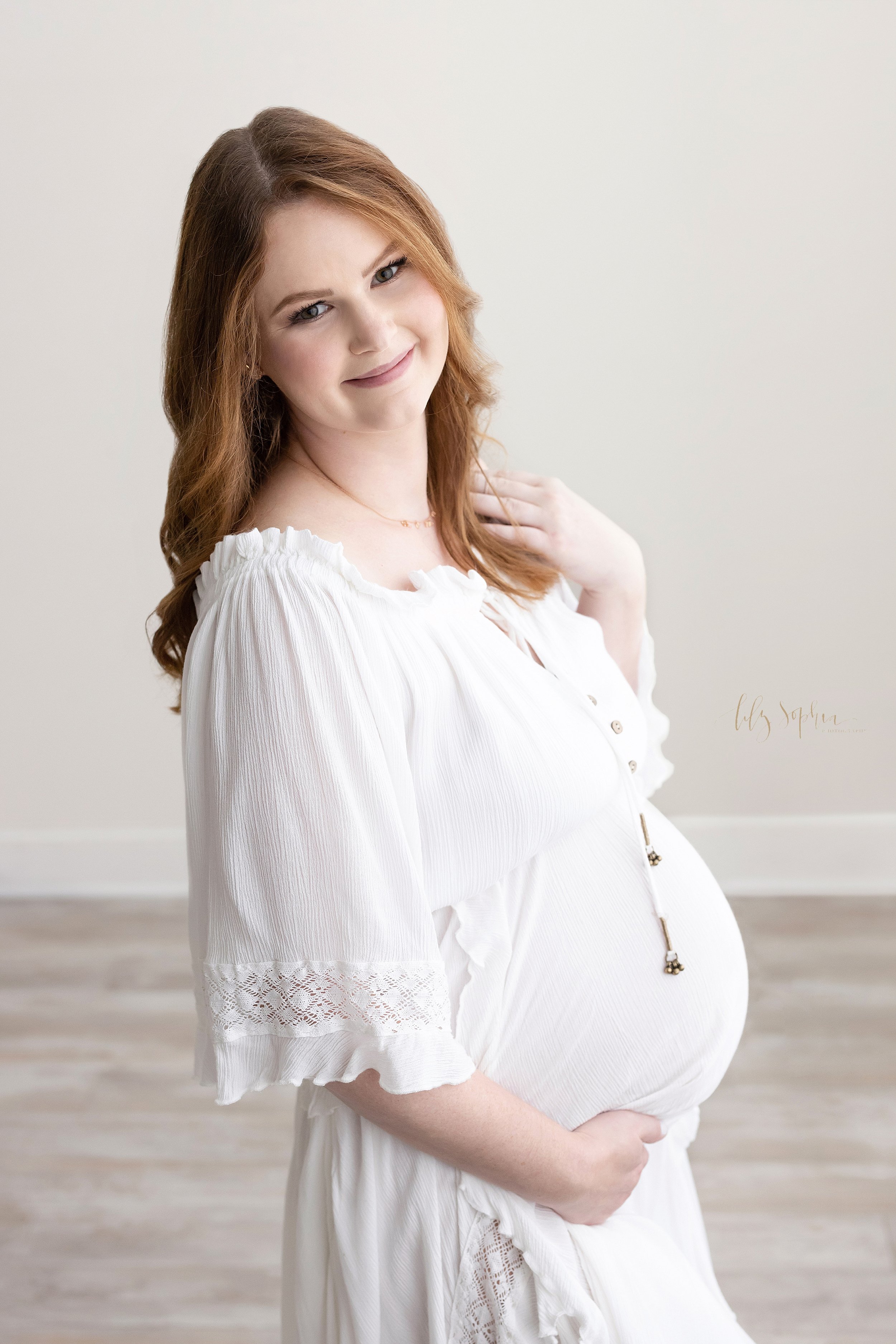  Maternity photo shoot of a red-headed pregnant mother standing in a natural light studio looking over her right shoulder with her right hand framing her belly and her left hand stroking her hair taken near Oakhurst in Atlanta. 