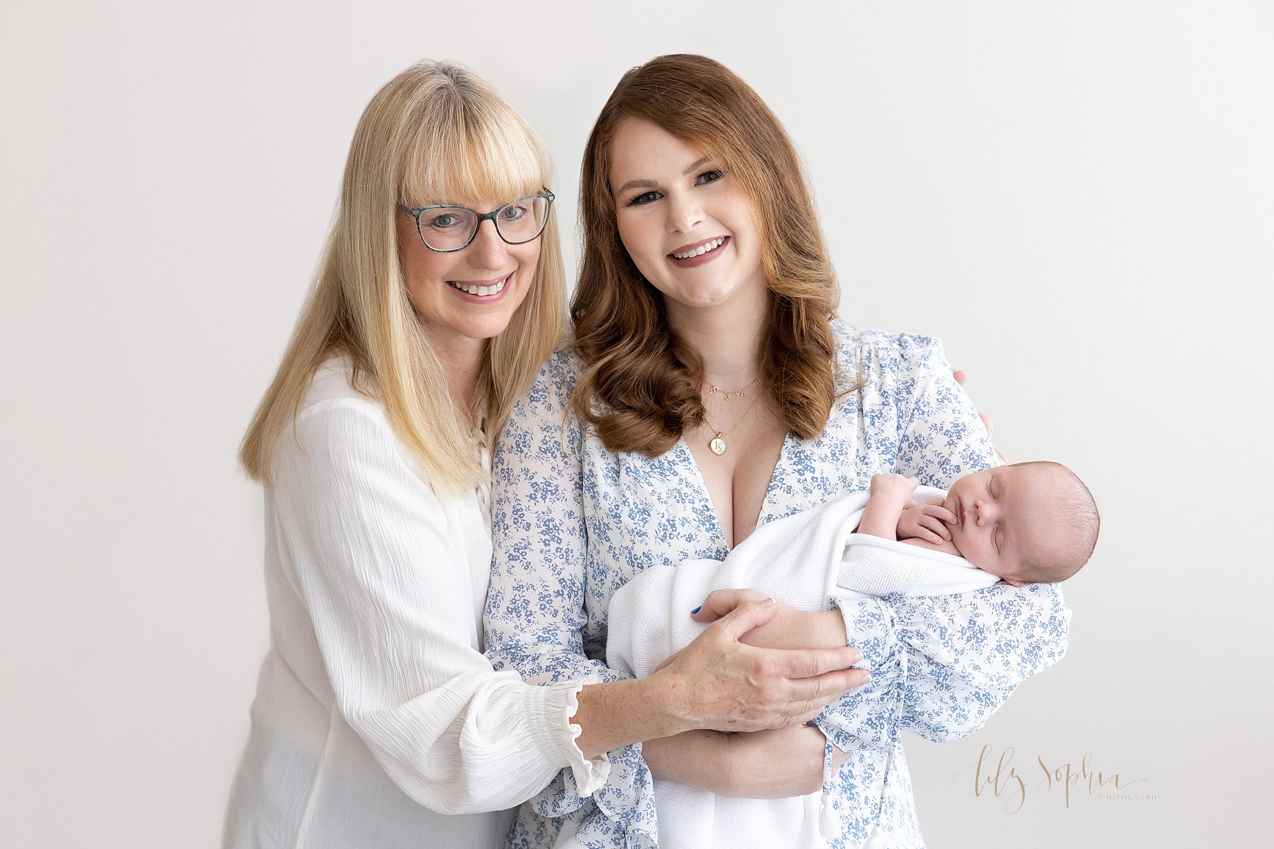  Intergenerational newborn photo of a mother holding her newborn son as the grandmother places her hand on her daughter’s while they stand in front of a window streaming natural light in a studio near Oakhurst in Atlanta. 