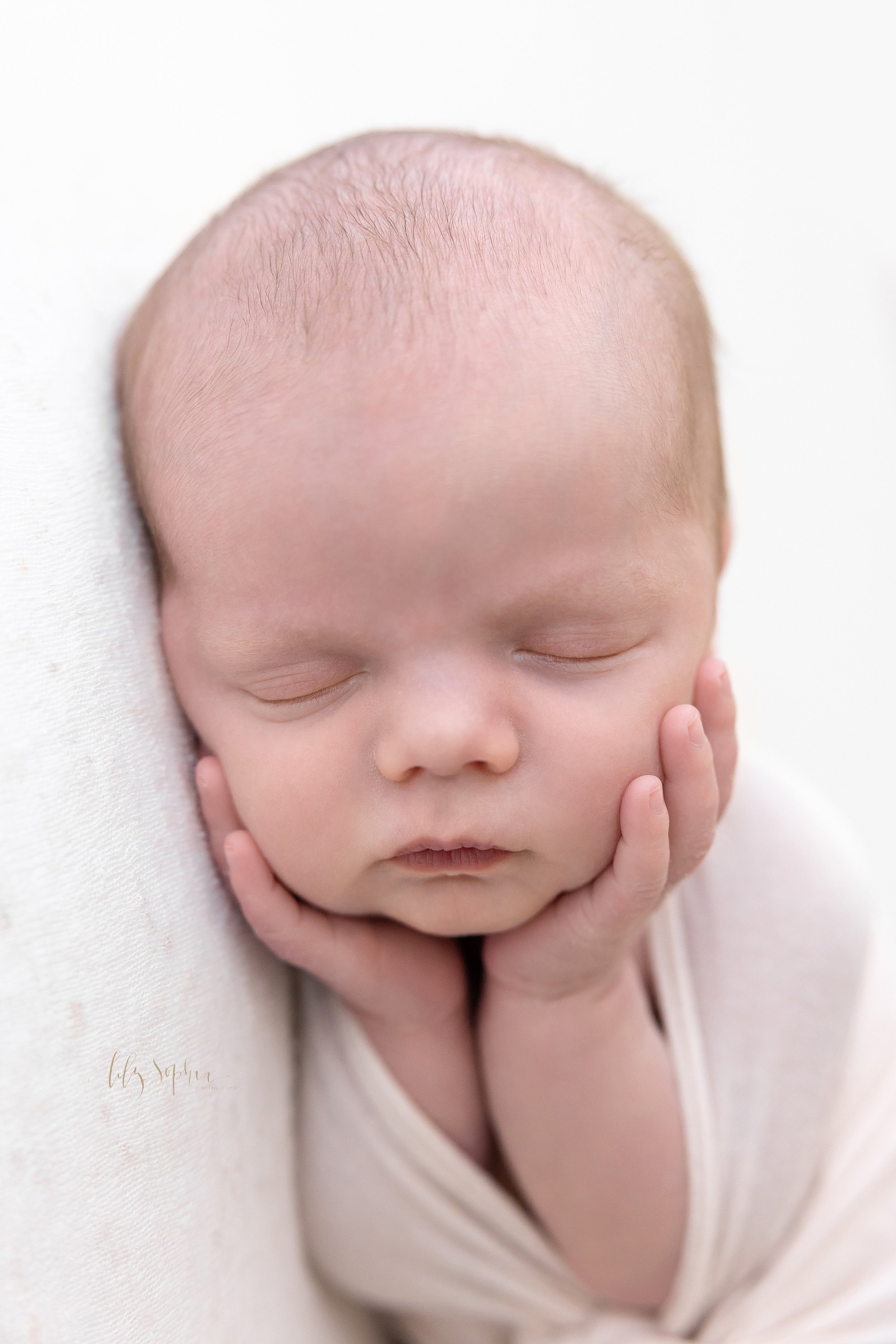  Close-up newborn portrait of a peacefully sleeping infant boy with his hands holding his cheeks taken in a natural light studio near Roswell in Atlanta. 
