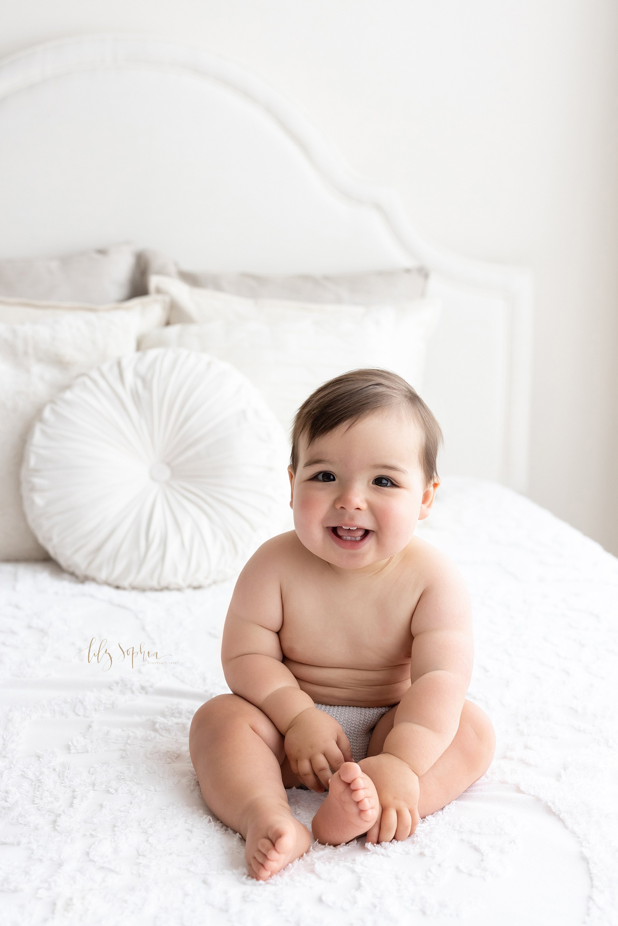  First birthday portrait of a smiling one year old boy as he sits in a diaper cover on top of a bed with his tiny teeth showing taken in a natural light studio near Midtown in Atlanta, Georgia. 