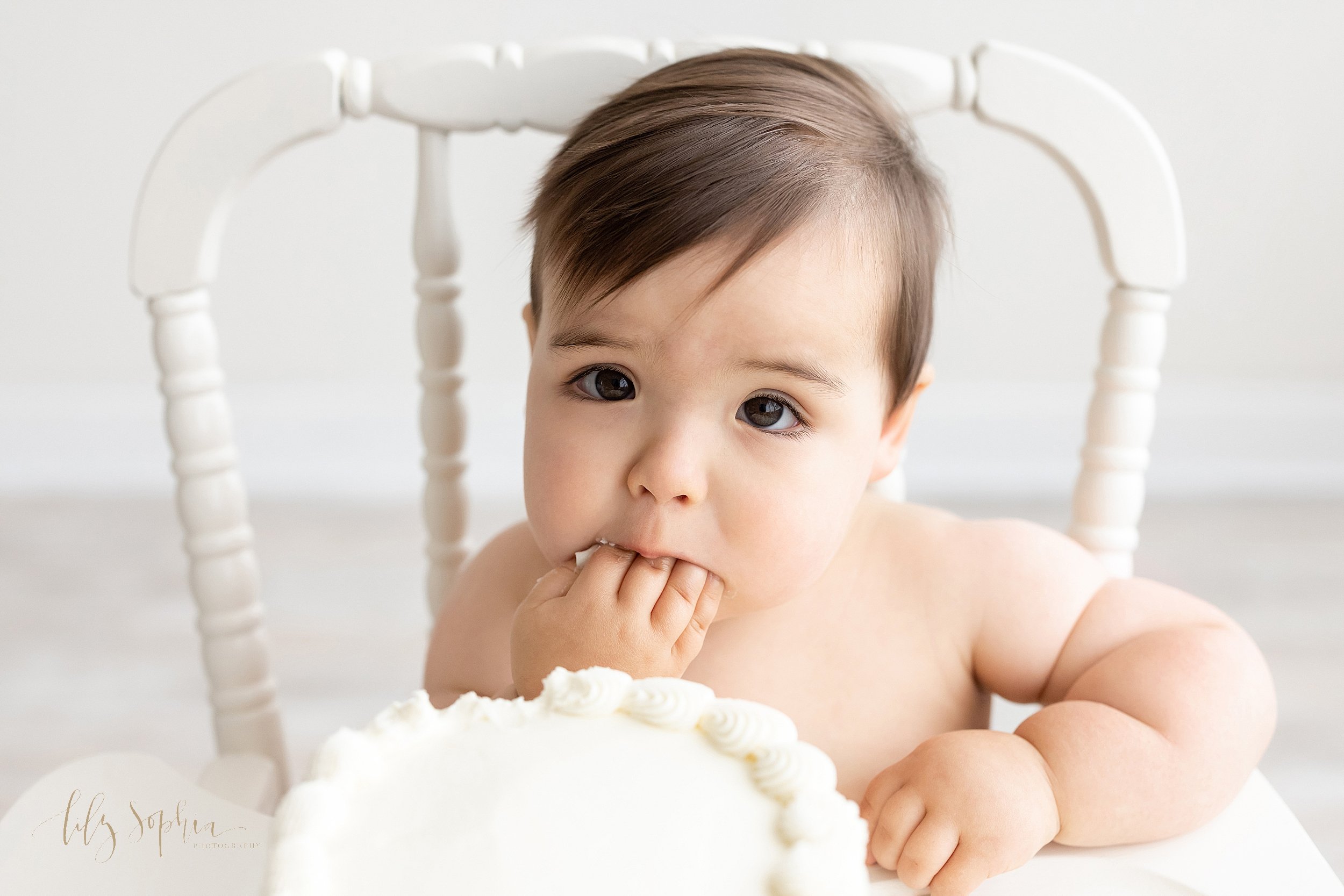  First birthday smash cake photo of a one year old little boy sitting in an antique high chair with his right hand shoving cake into his mouth taken in a studio near Alpharetta in Atlanta that uses natural light. 