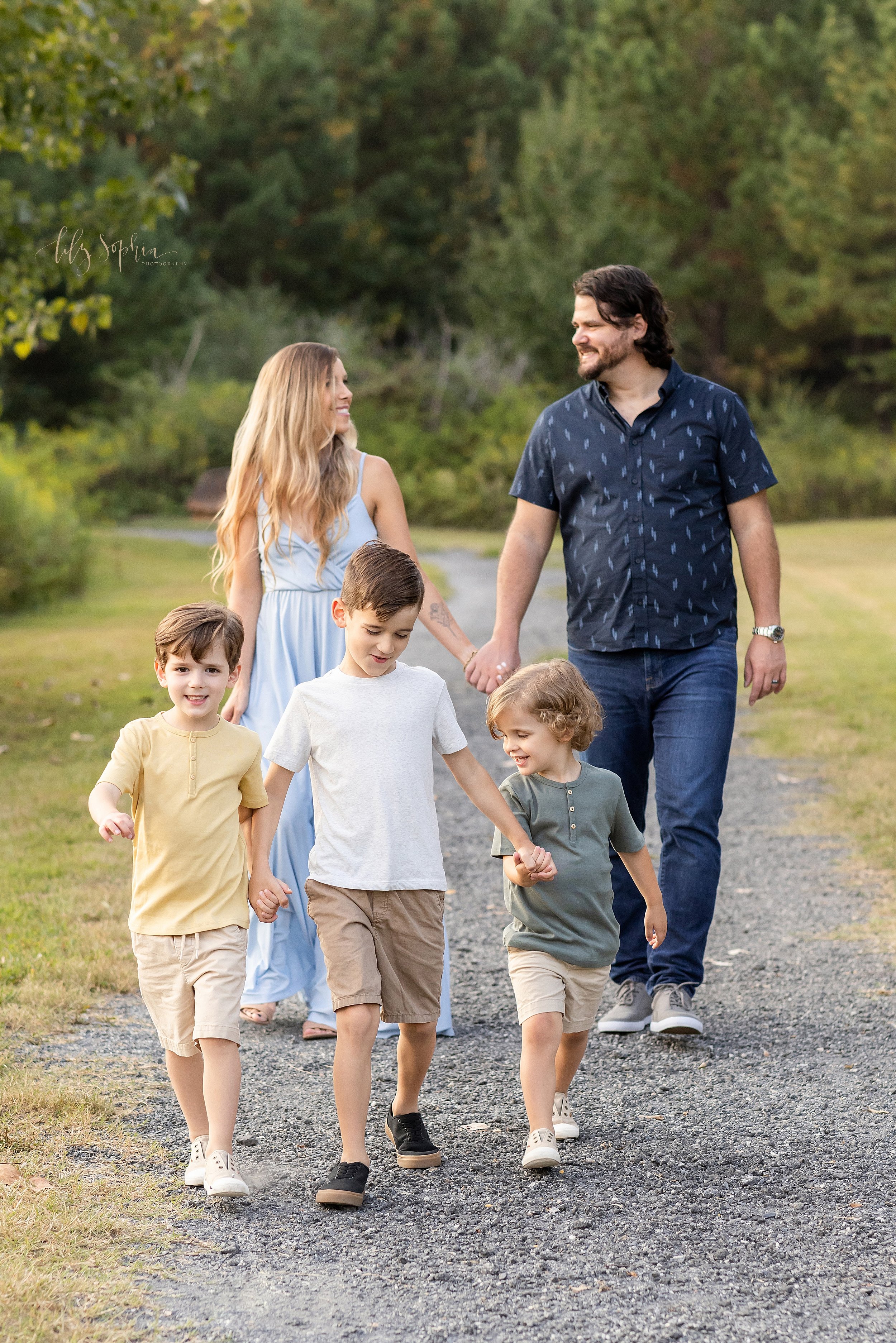  Family photo shoot of a mother and father walking hand-in-hand behind their three sons who are walking hand-in-hand on a path at sunset in an Atlanta park. 