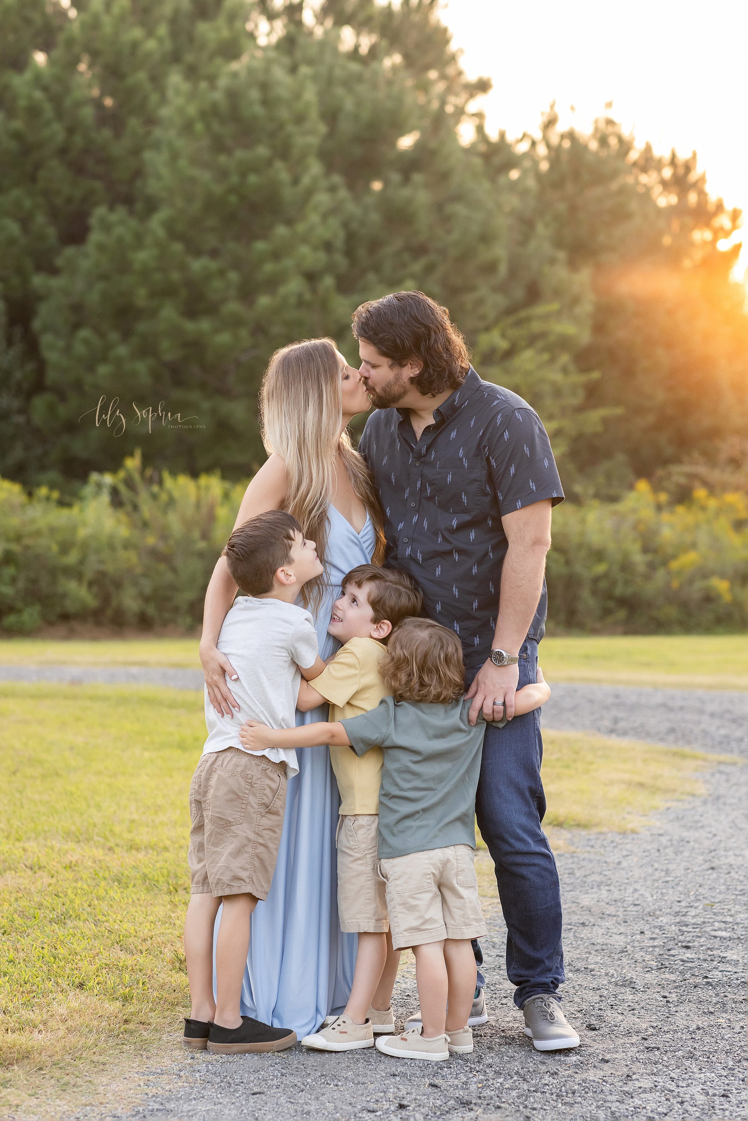  The love of a family is captured in this family portrait of a mother and father kissing one another as their three sons reach to hug them taken in a park in Atlanta at sunset. 