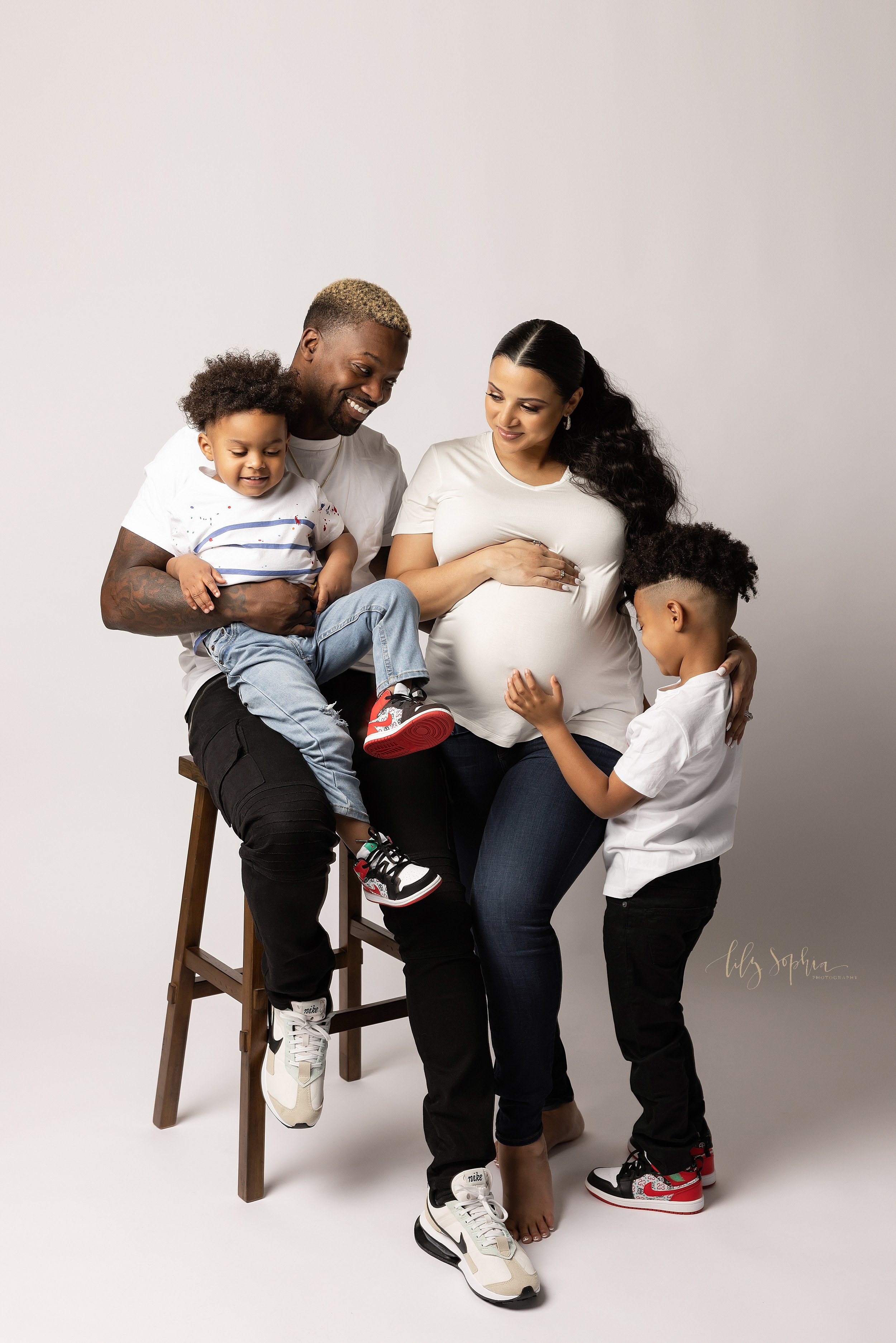  Family maternity portrait with dad sitting on a wooden stool and holding his young son on his lap as he sits next to his pregnant wife sitting on another stool with her right hand on top of her belly and her left hand on her older son’s shoulder as 