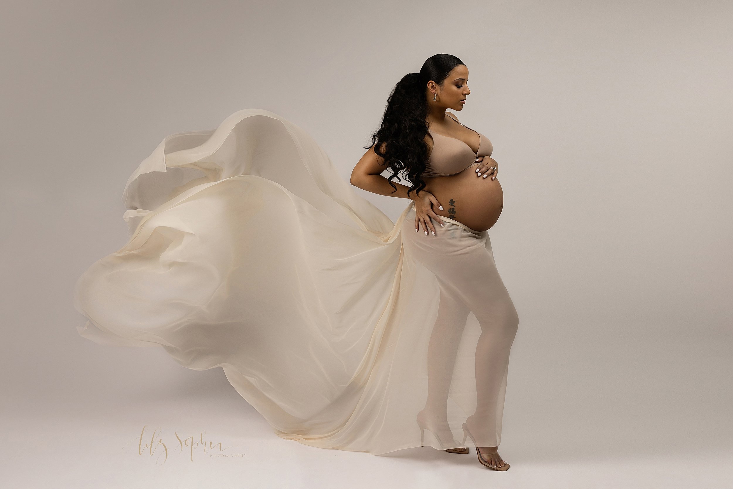  Modern maternity photo session with a pregnant woman wearing a full-length billowing sheer skirt and a bra with her belly bare and her right hand on her hip and her left hand on top of her belly to show her profile taken in a studio near Old Fourth 