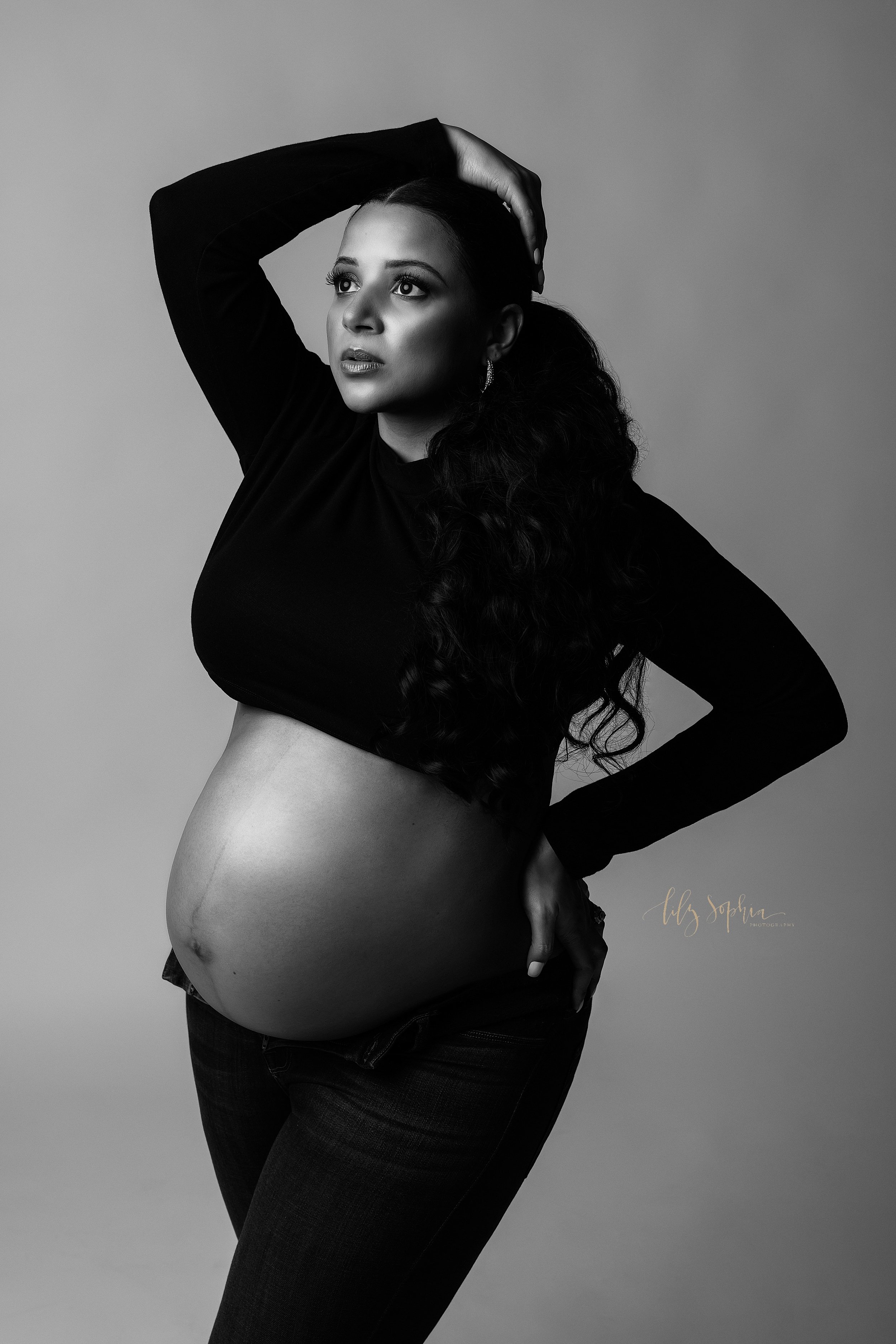  Modern maternity black and white portrait of a pregnant woman wearing unzipped jeans ad a long sleeved midriff knit pull-over with her right hand on top of her head and her left hand on her left hip taken in a studio near Kirkwood in Atlanta. 