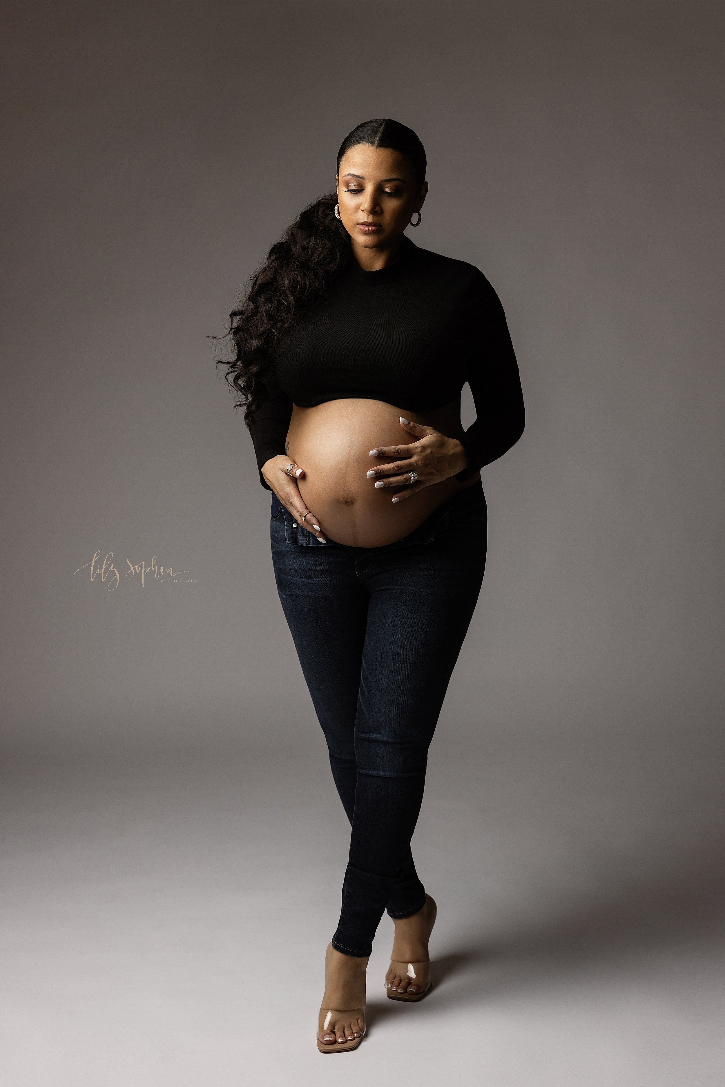  Modern maternity photo of an a woman wearing unzipped blue jeans and a black knit long-sleeved midriff pull-over with her belly bare and her hands caressing her baby in utero taken in a natural light studio in Ponce City Market in Atlanta, Georgia. 