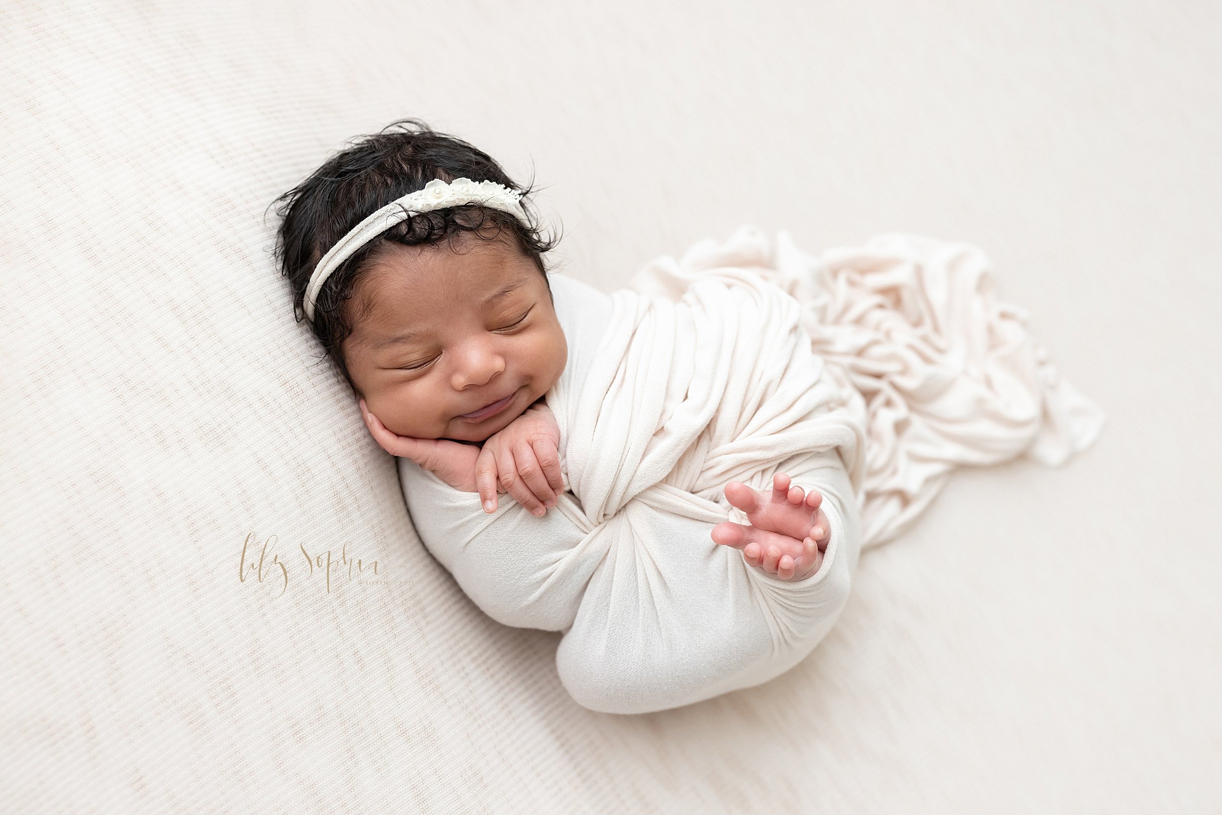  Newborn photo of a baby girl cradled in a stretchy swaddle with her tiny toes peeking out and her right hand holding her cheek and her left hand under her chin as she smiles in her sleep wearing a stretchy rose headband in her curly black hair taken